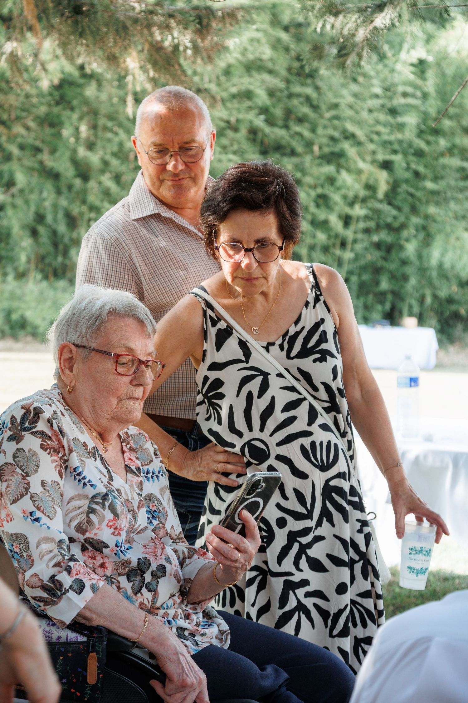 Elderly woman in wheelchair views phone held by younger woman while man stands behind at outdoor event.