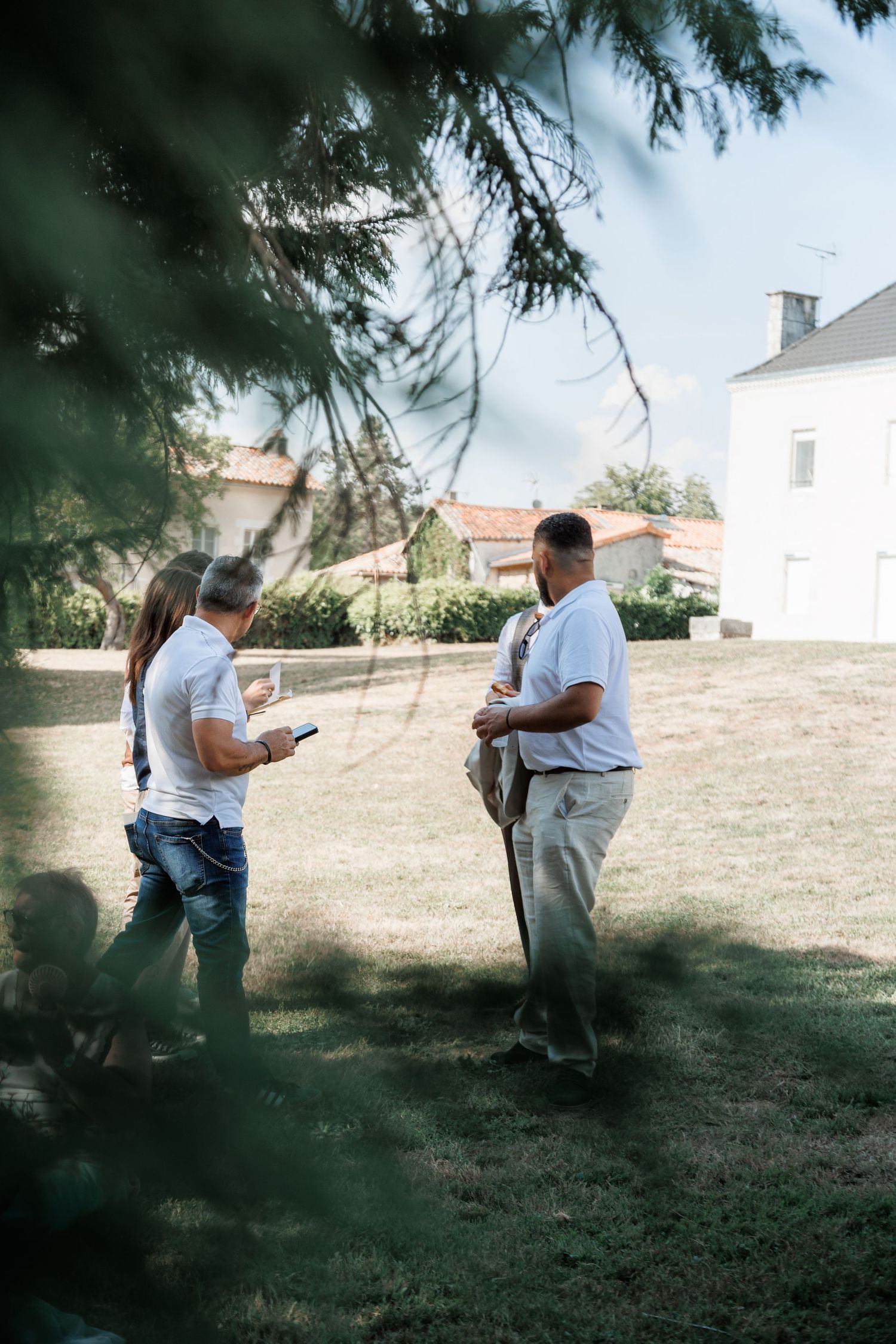 Three guests chat on sunny lawn outside white country house during outdoor wedding reception.