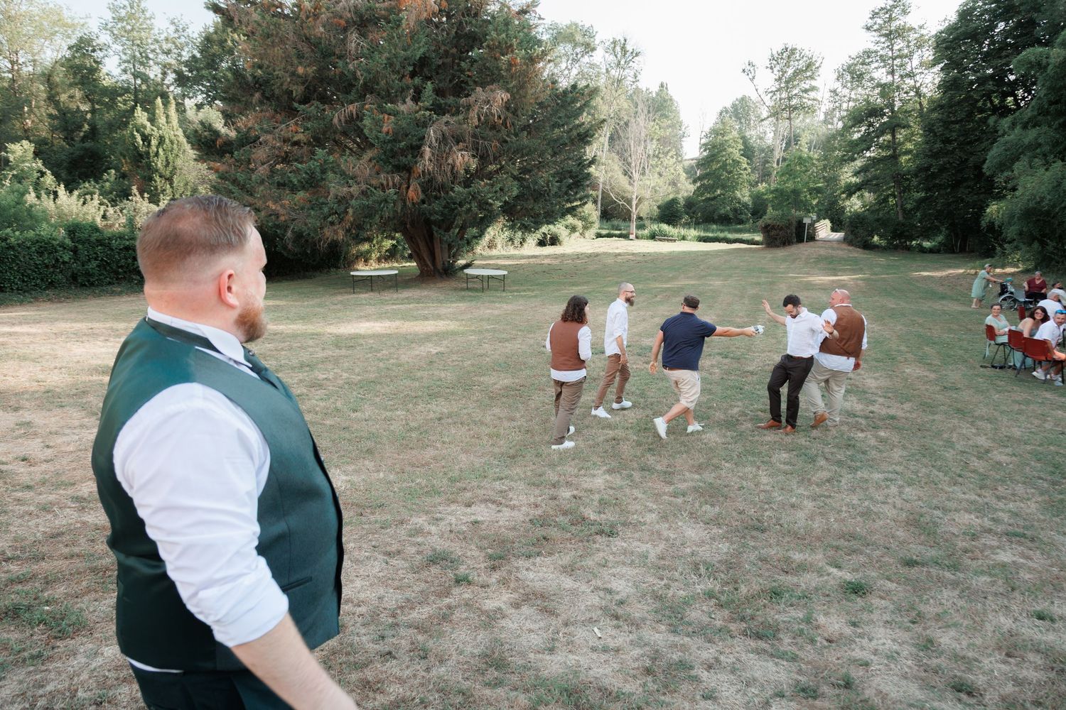 Groom in green vest watching wedding guests play lawn games in sunny outdoor garden reception venue.