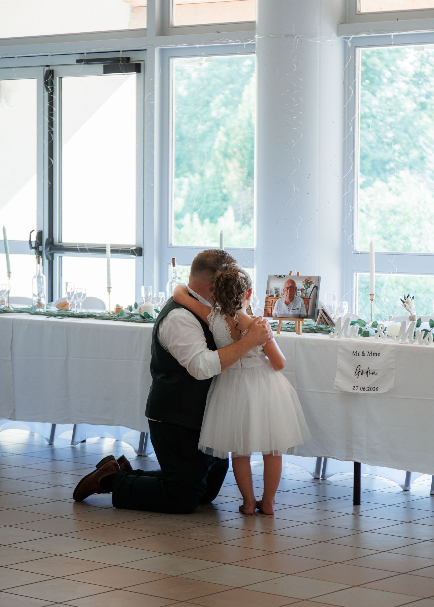 Groom kneeling to embrace flower girl in white tutu dress at wedding reception venue.