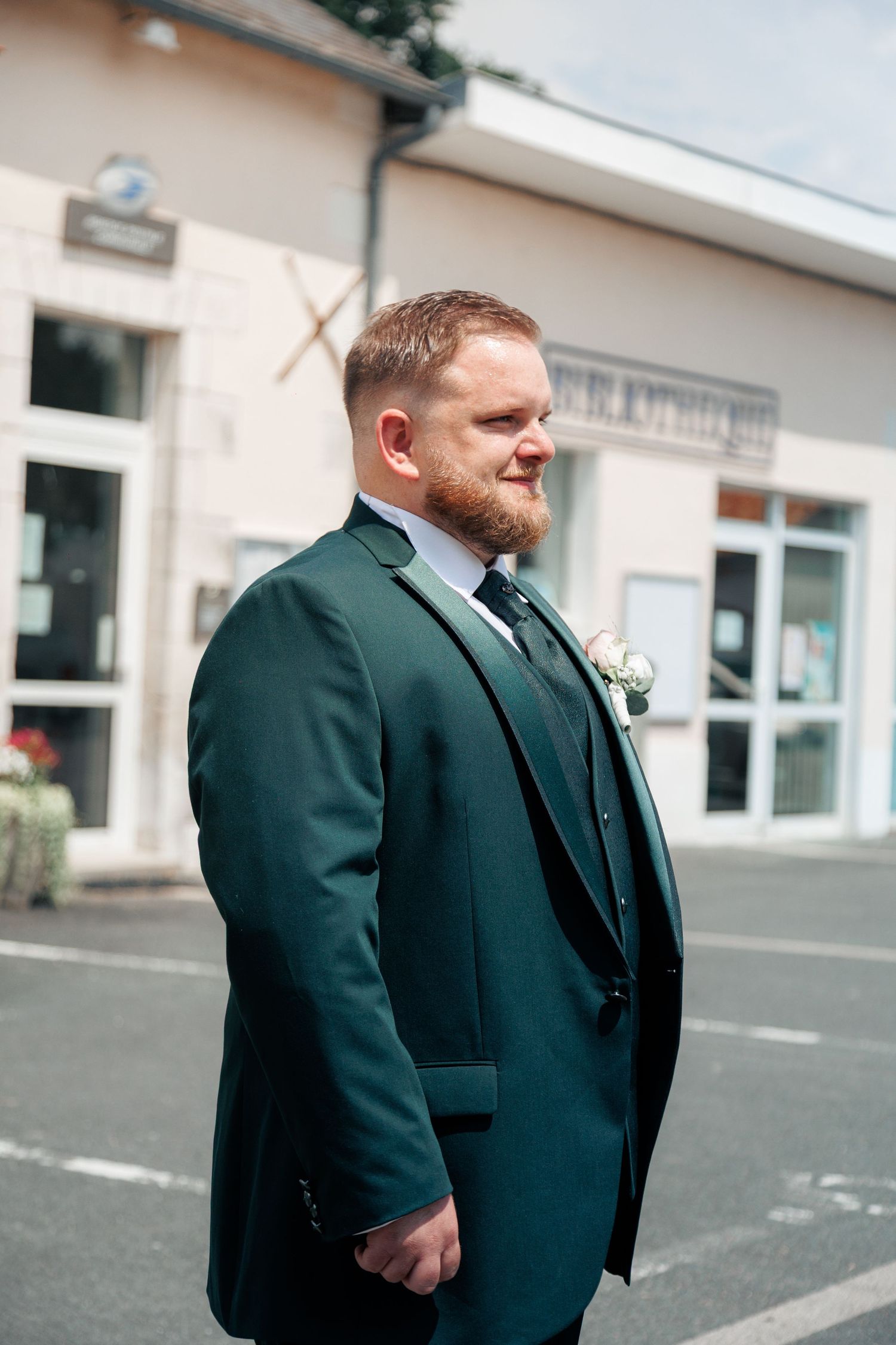 Handsome groom in dark green suit with boutonniere standing outdoors before wedding ceremony.