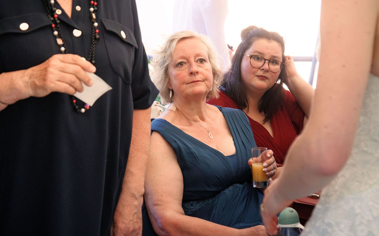 Elegant wedding guests in blue and burgundy dresses attend indoor ceremony, one holding orange drink.