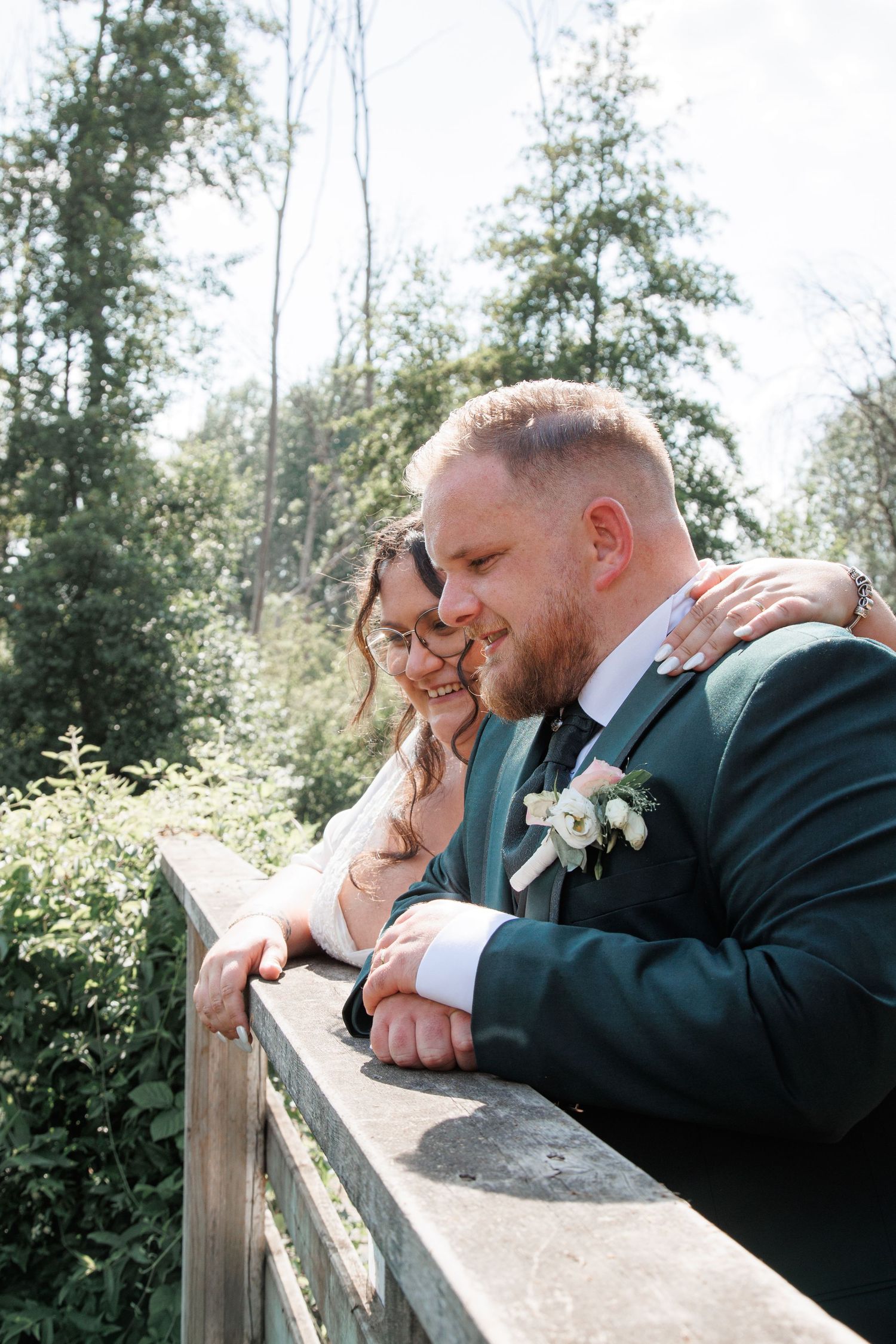 Bride and groom leaning on wooden bridge railing in forest setting.
