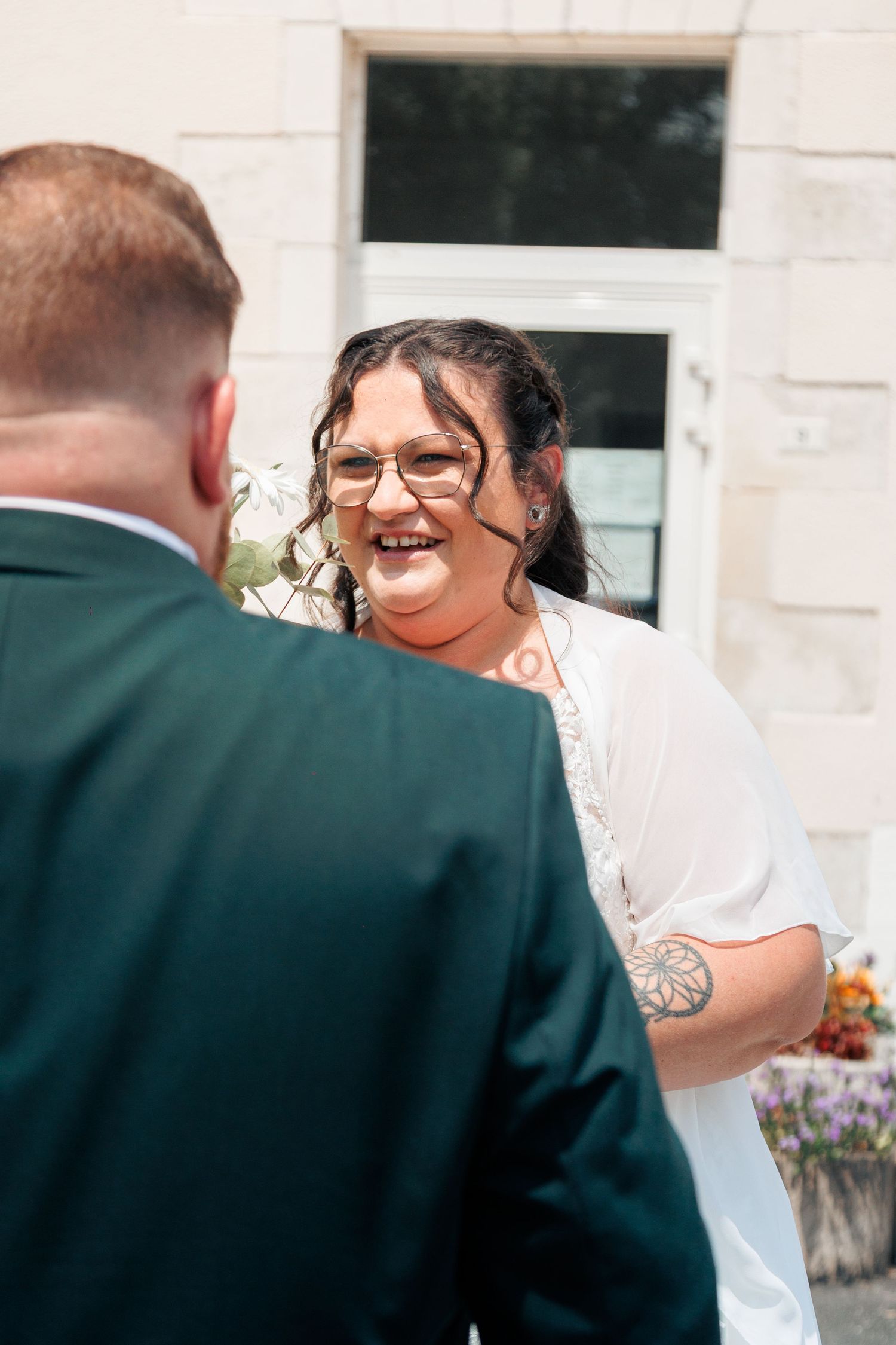 Smiling bride in white dress and glasses sharing a joyful moment with groom in dark green suit outdoors.