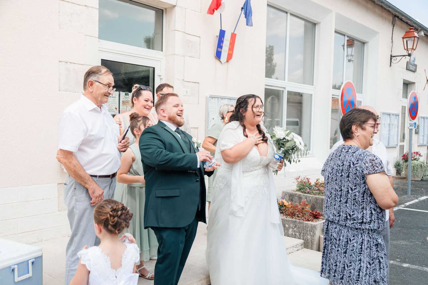 Wedding couple in white dress and green suit celebrating outside French town hall with family guests.