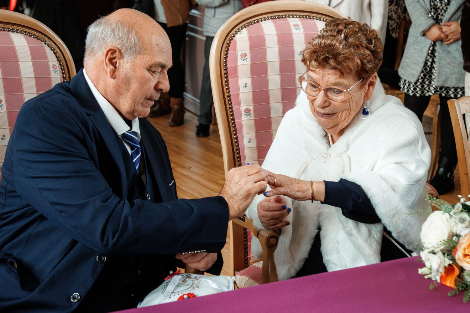 Elderly couple exchanging rings at a wedding ceremony, seated at a decorated table with flowers.