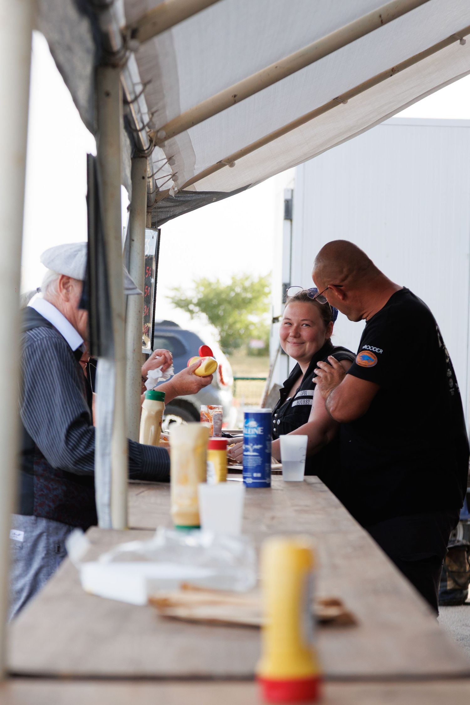 People enjoying food and drinks at an outdoor food stand, smiling and socializing at a wooden counter.