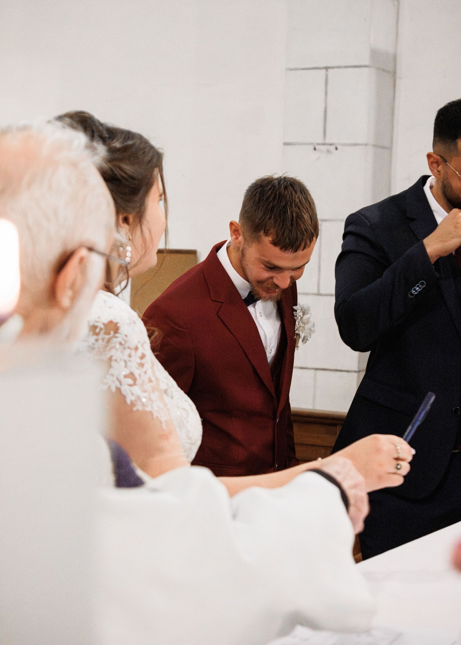 Groom in burgundy suit bowing head during wedding signing ceremony beside bride in lace dress.