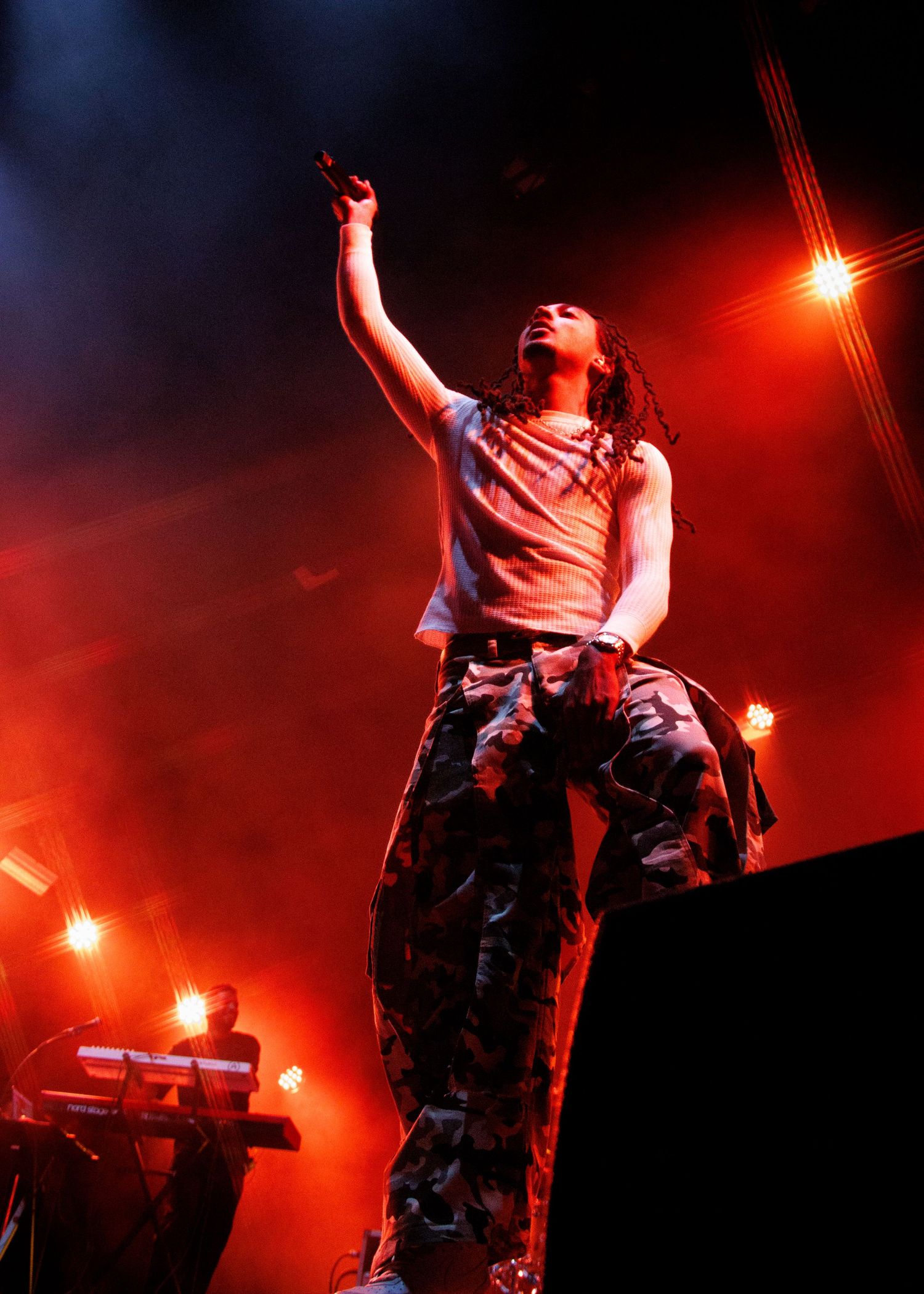 A performer in camo pants raises their arm dramatically on a smoky, red-lit concert stage.