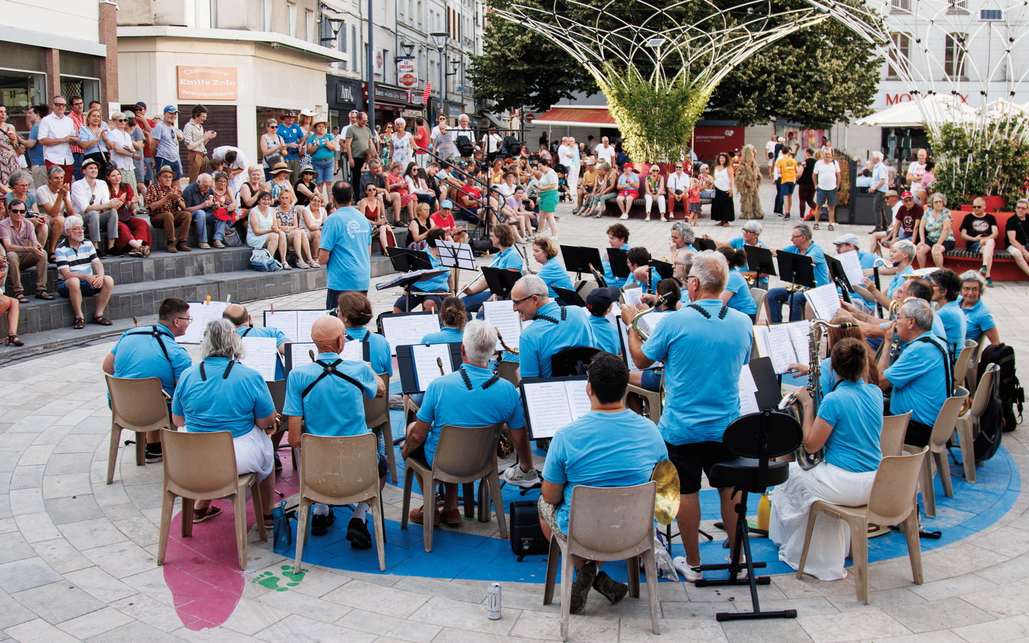An outdoor concert featuring musicians in blue shirts performing in a crowded plaza.