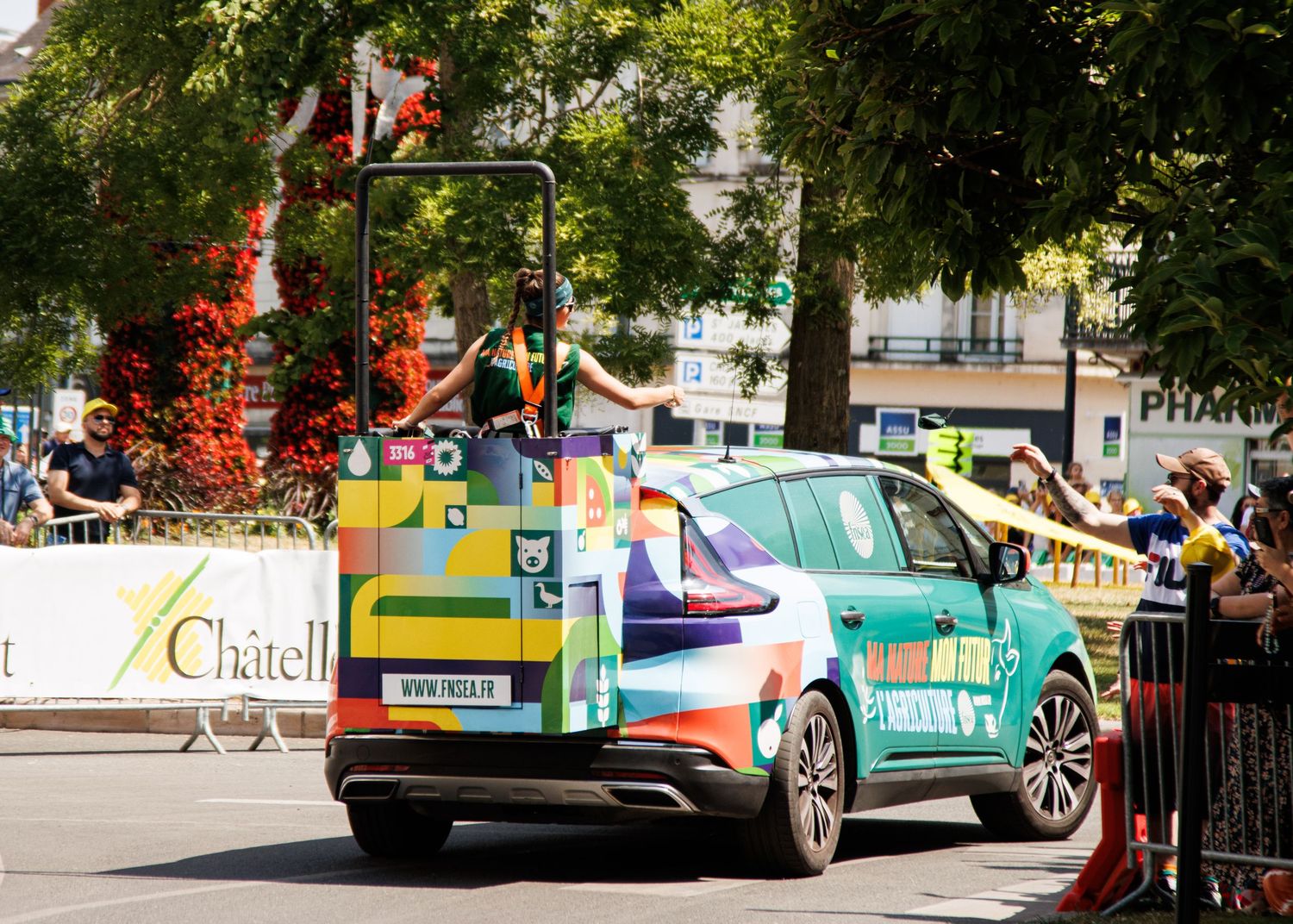 Colorful branded vehicle participates in parade procession on tree-lined urban street.