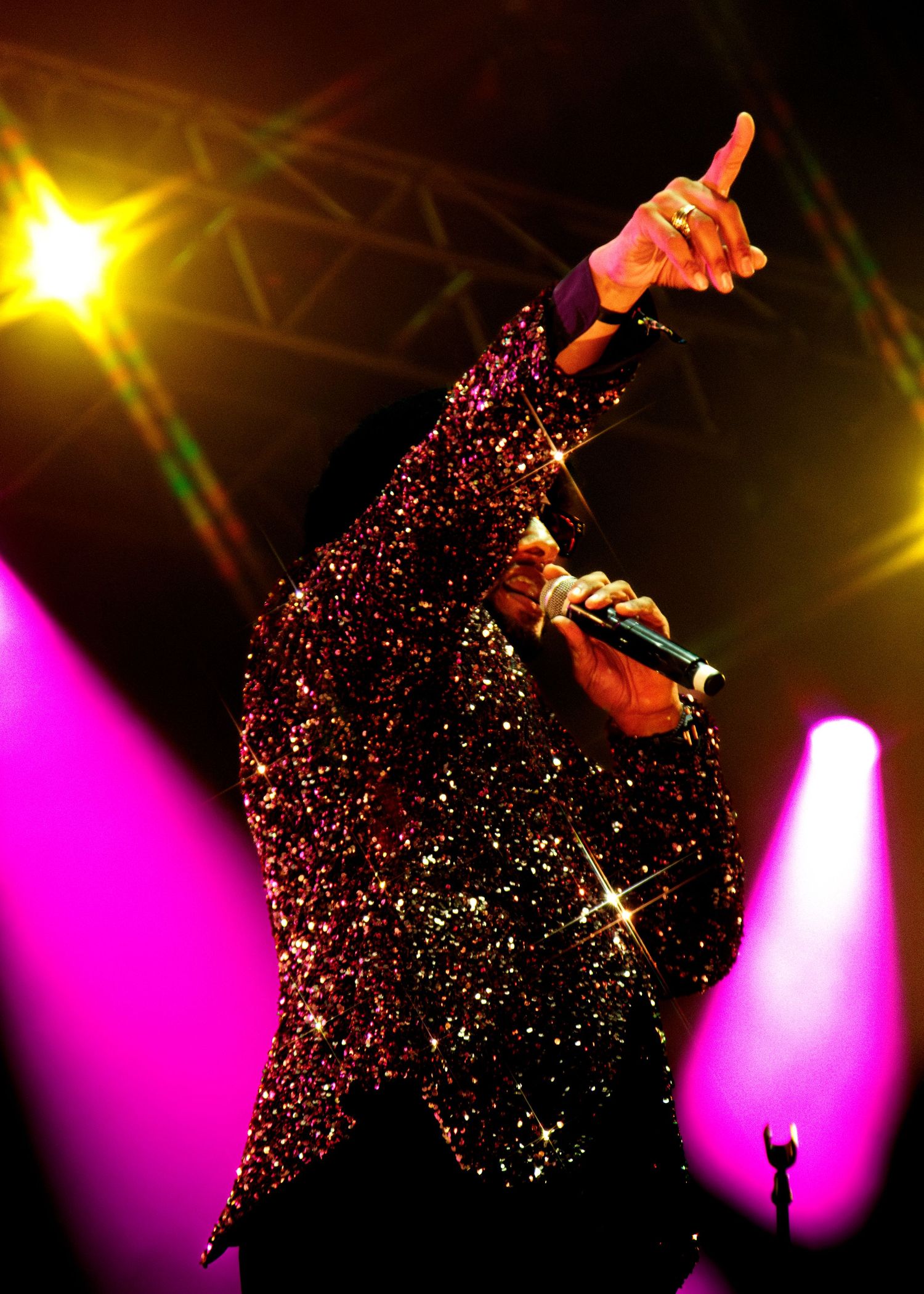 Performer in sparkling black outfit raises arm while singing on stage with colorful purple and yellow concert lighting.
