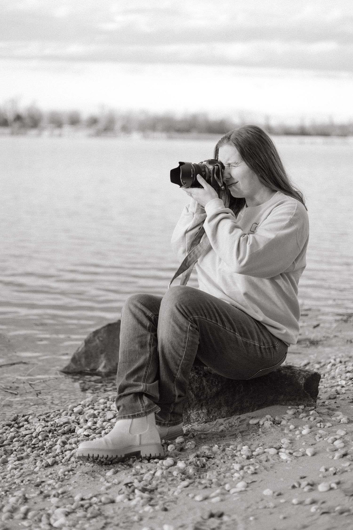 A photographer in casual attire lies on a rocky beach taking pictures with a telephoto lens near the water's edge.