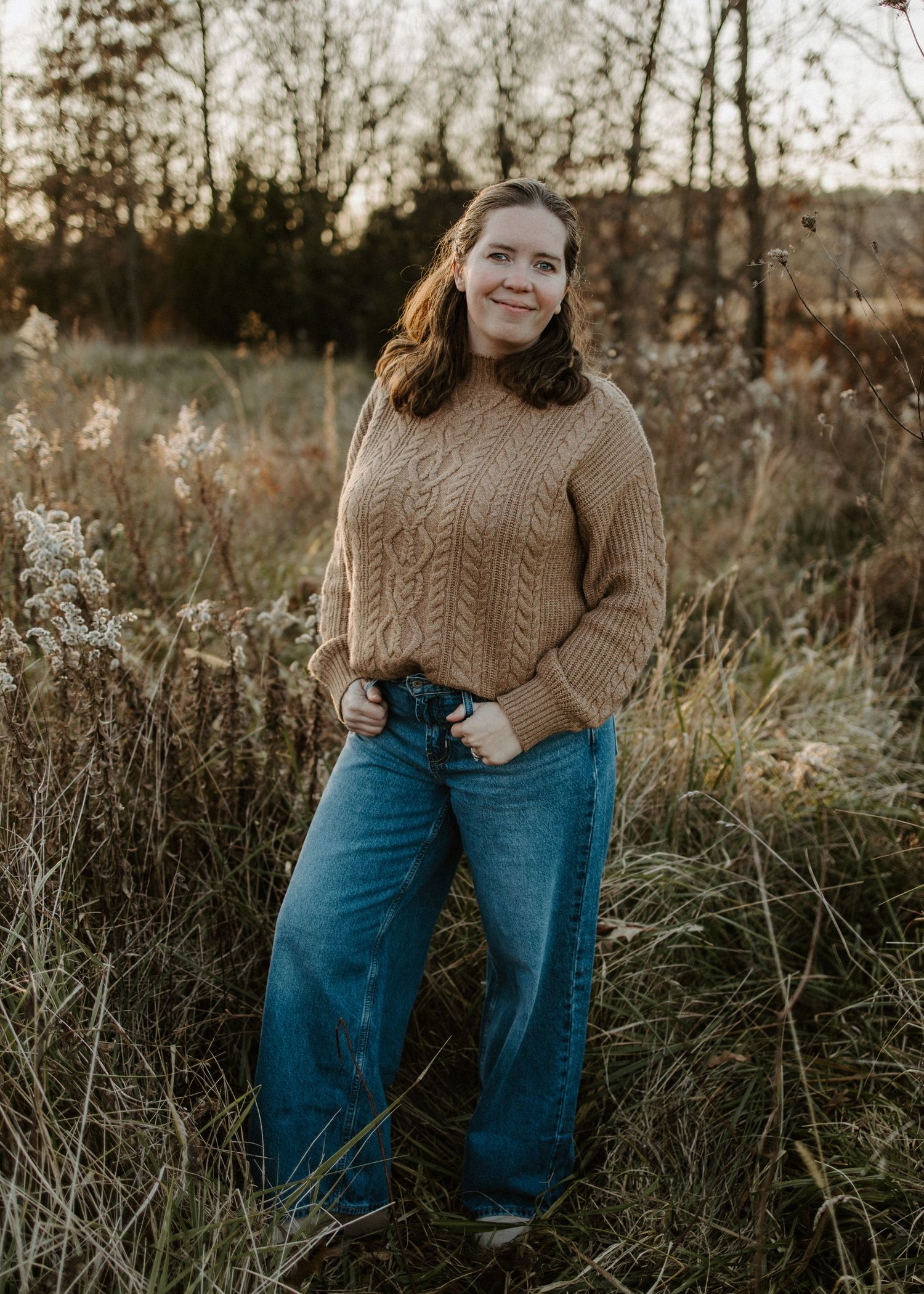A figure in a cozy brown cable knit sweater and blue jeans poses casually in a field of tall grass at sunset.