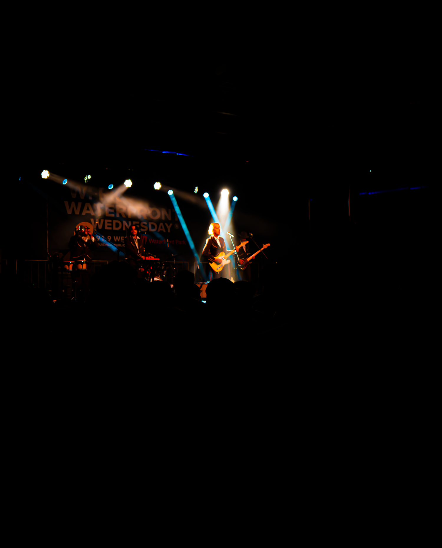 Silhouetted musicians perform on a dark stage with dramatic red and blue lighting effects in the background.