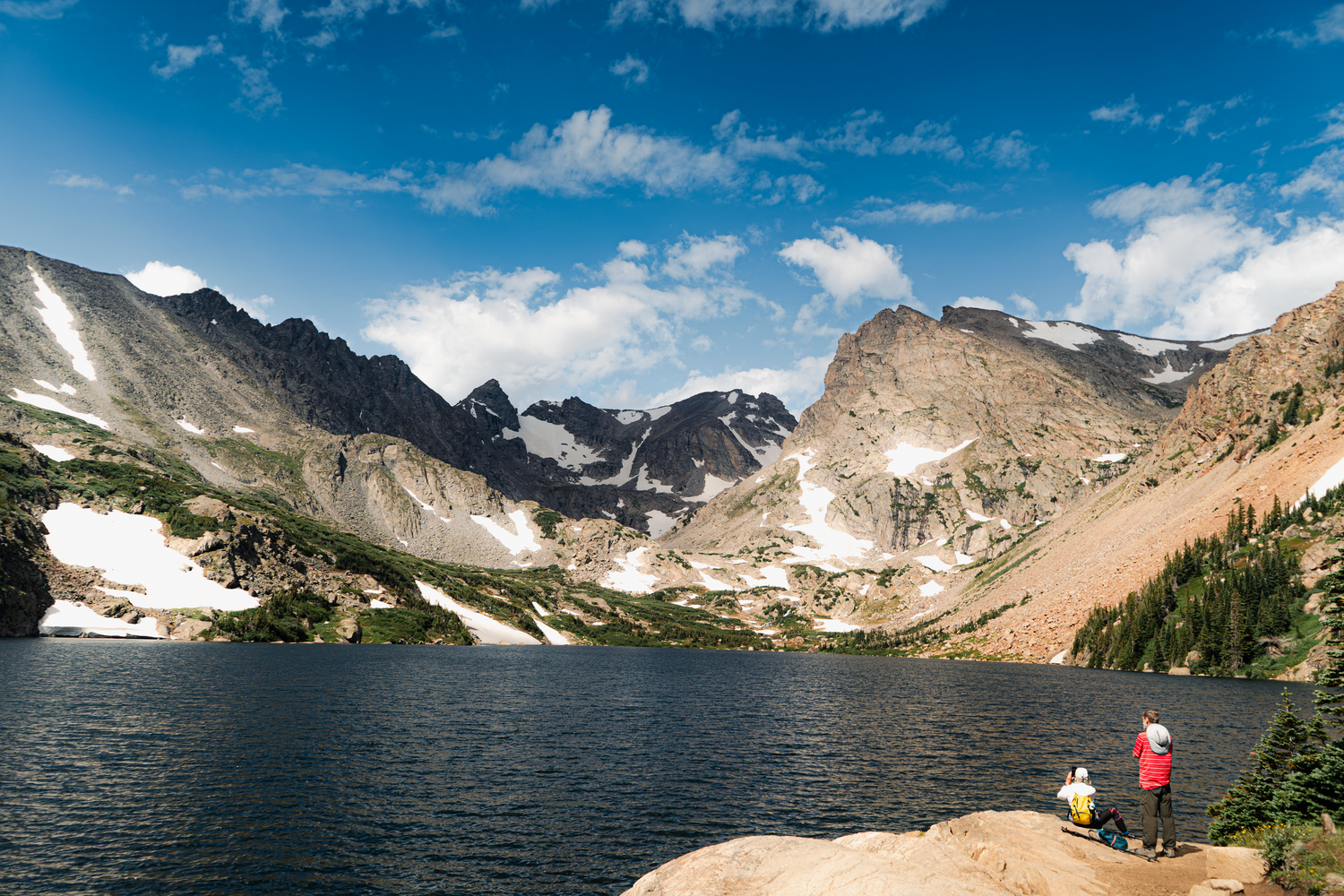 A hiker admires the scenic mountain landscape with snow patches and alpine lake under blue skies in Indian Peaks Wilderness.