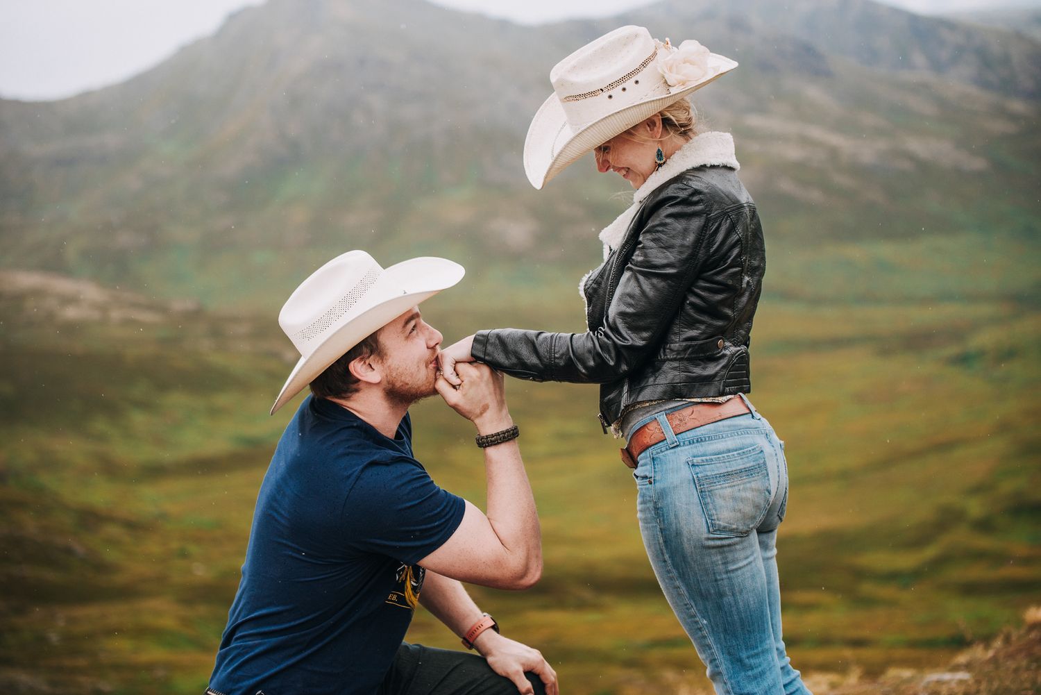 Romantic proposal scene with cowboy hats in mountain landscape.