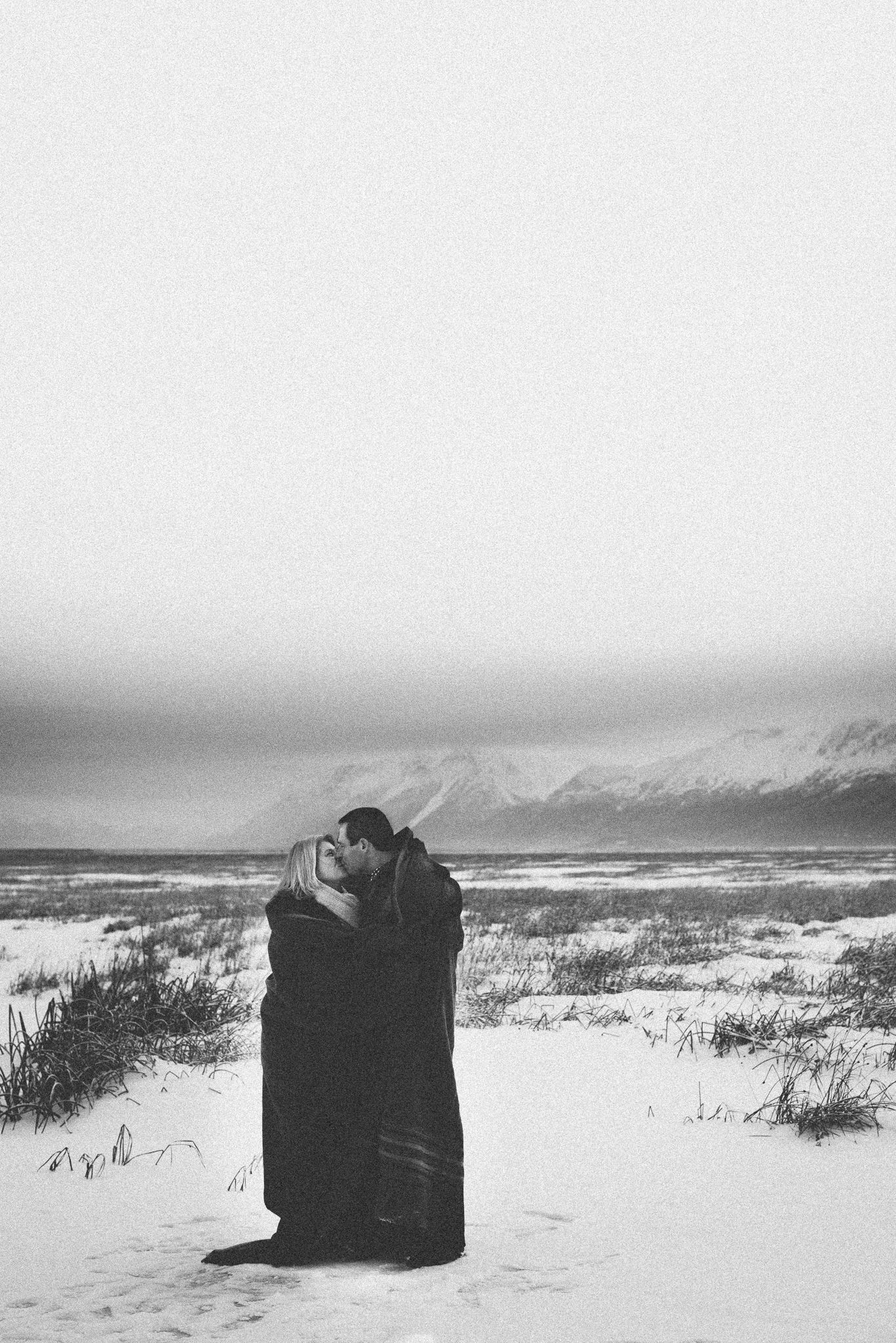Black and white photo of a couple embracing on a snowy beach with mountains in the distance.