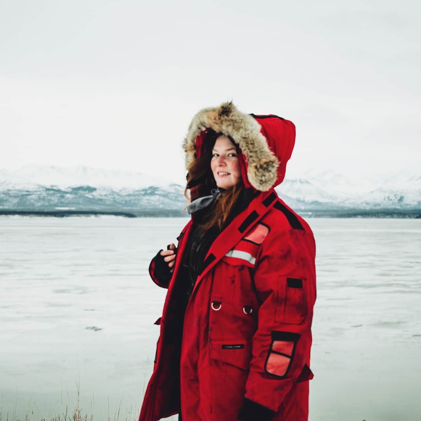Person wearing a fur-trimmed red winter parka poses against a wintry lake landscape.