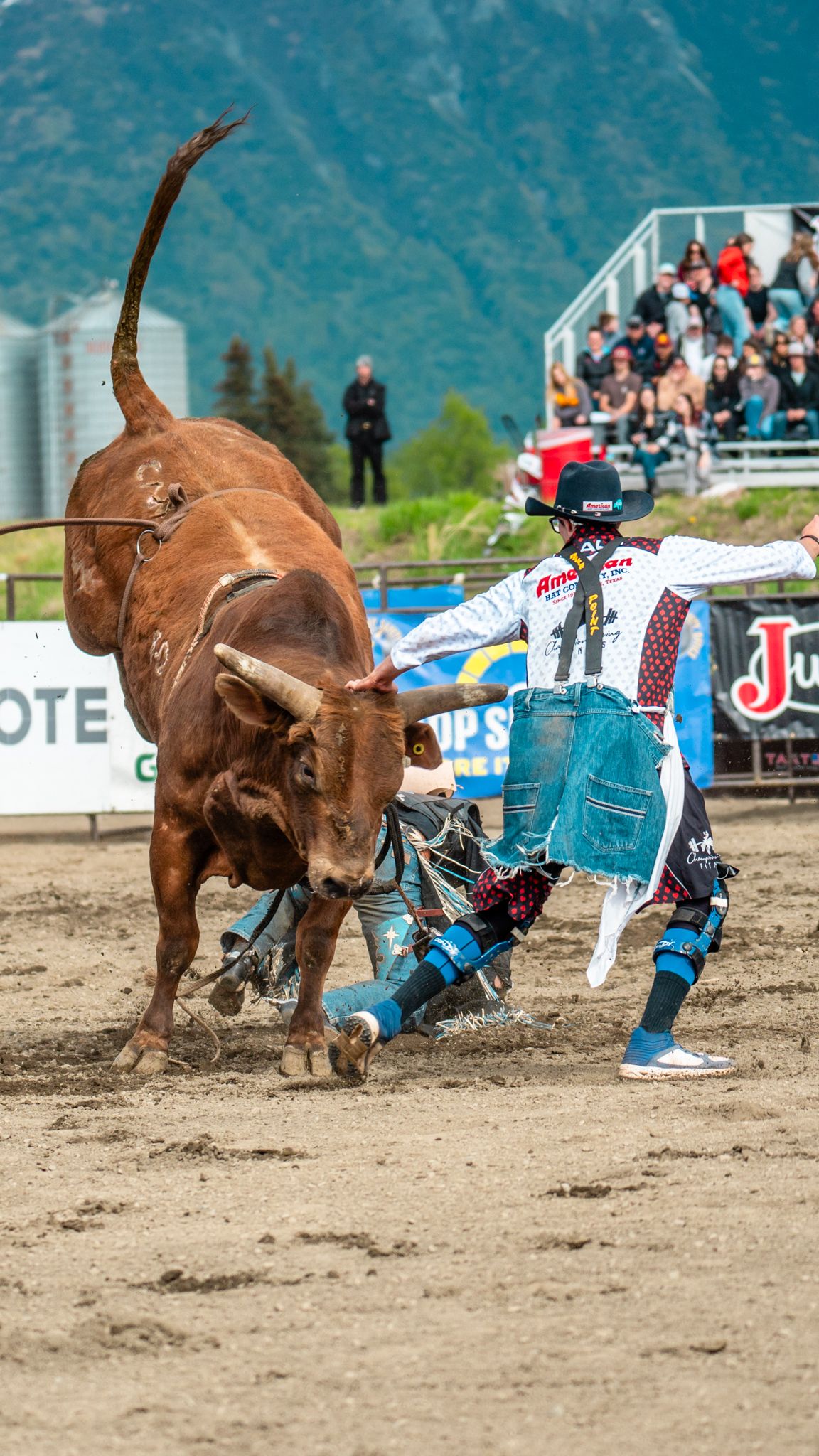 Bull rider gets thrown during intense rodeo competition with dramatic mountain backdrop.