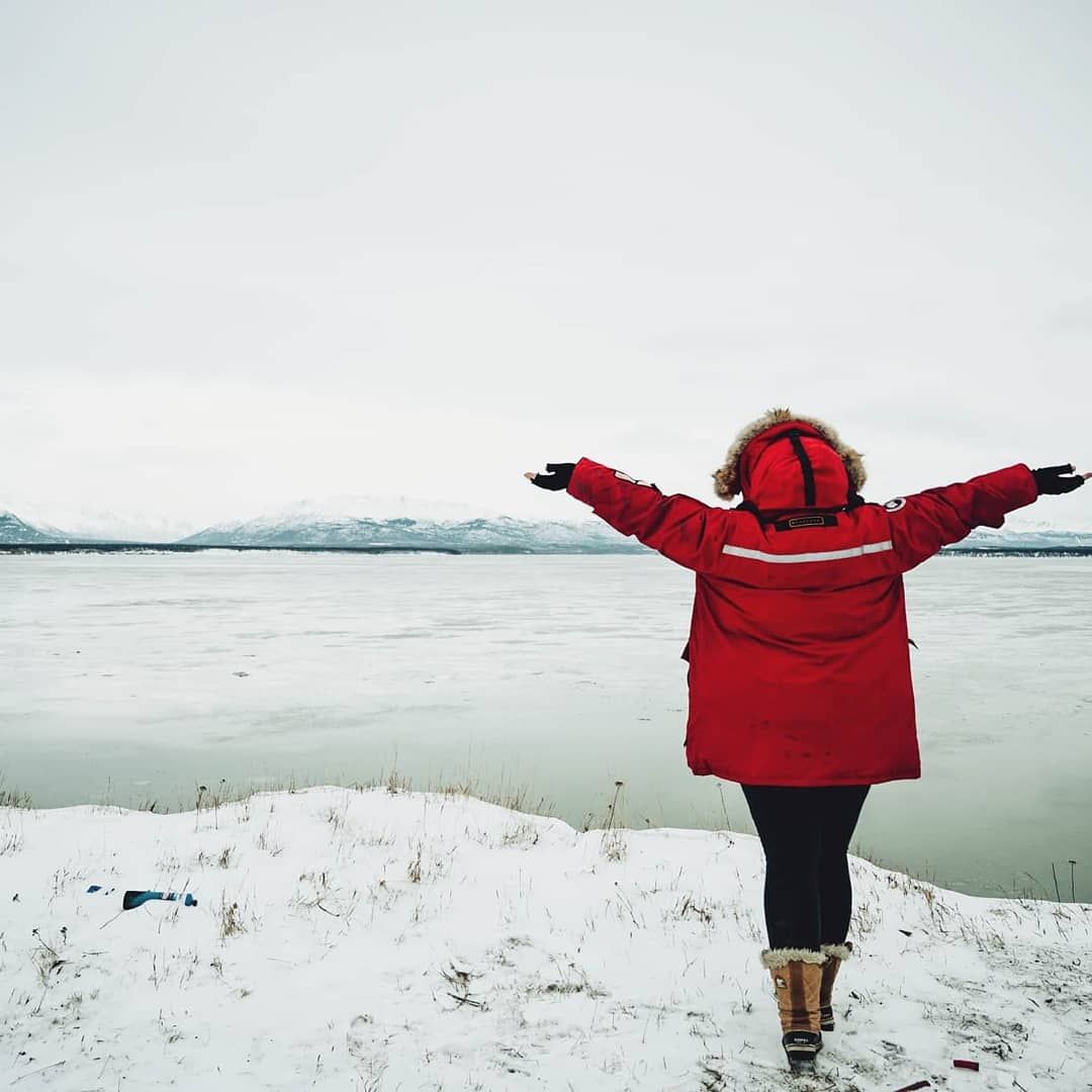 Person in red winter coat stands with arms outstretched on snowy shoreline overlooking frozen lake and mountains.