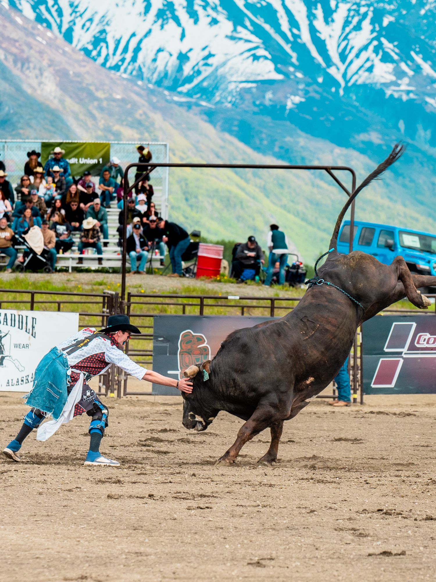 Dramatic bull riding action shot at an outdoor rodeo with mountain scenery.