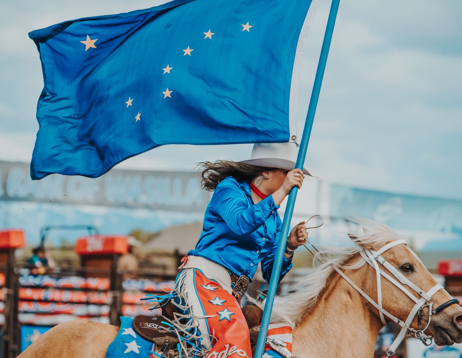 Rider carrying Alaska state flag on horseback during rodeo event.