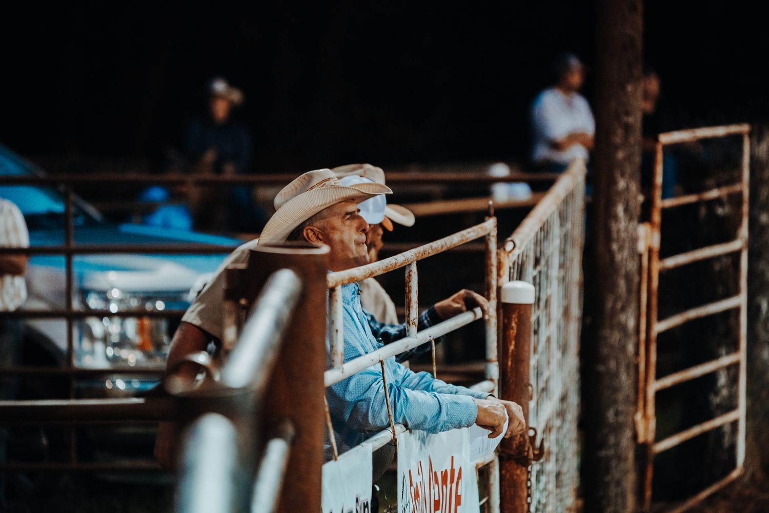 Cowboy leans against metal arena gates during evening rodeo event.