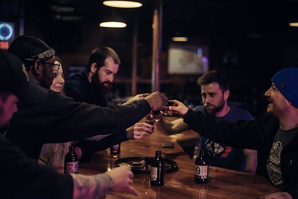 Friends toasting and clinking beer bottles together at a dark wooden bar.