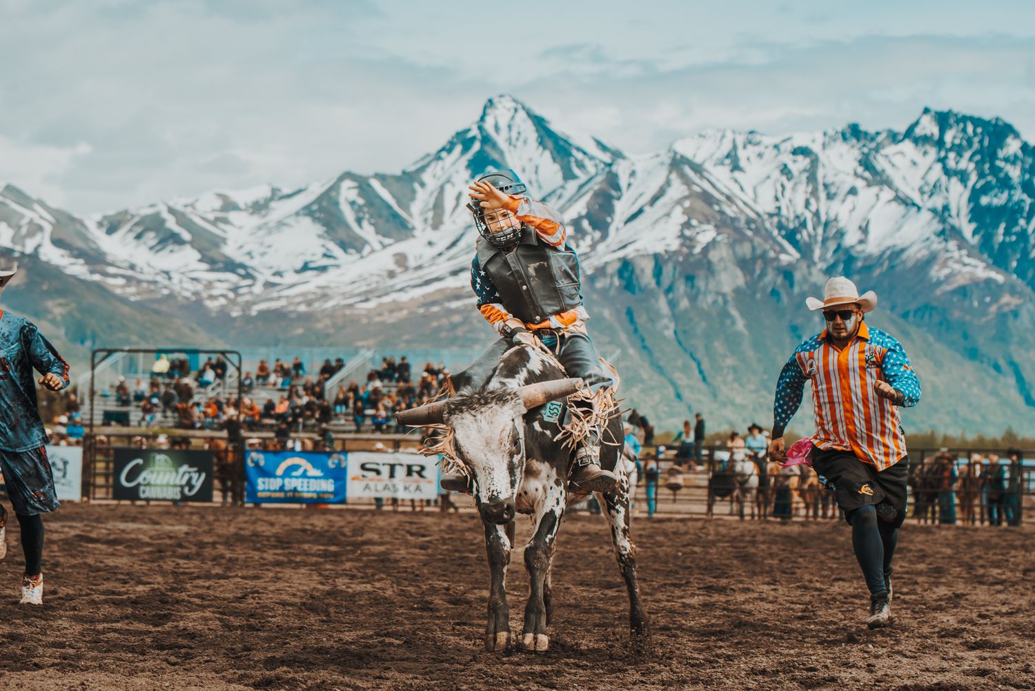 Majestic snow-capped mountain peaks tower over a rustic rodeo arena during a western competition event.