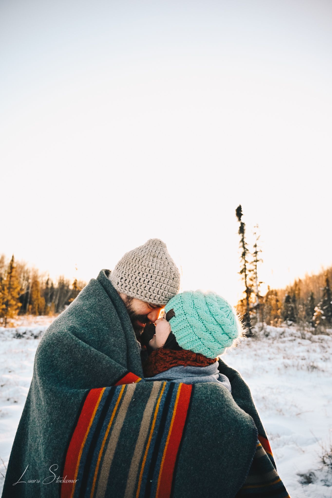Couple wrapped in a striped blanket sharing an intimate moment in a snowy winter landscape.