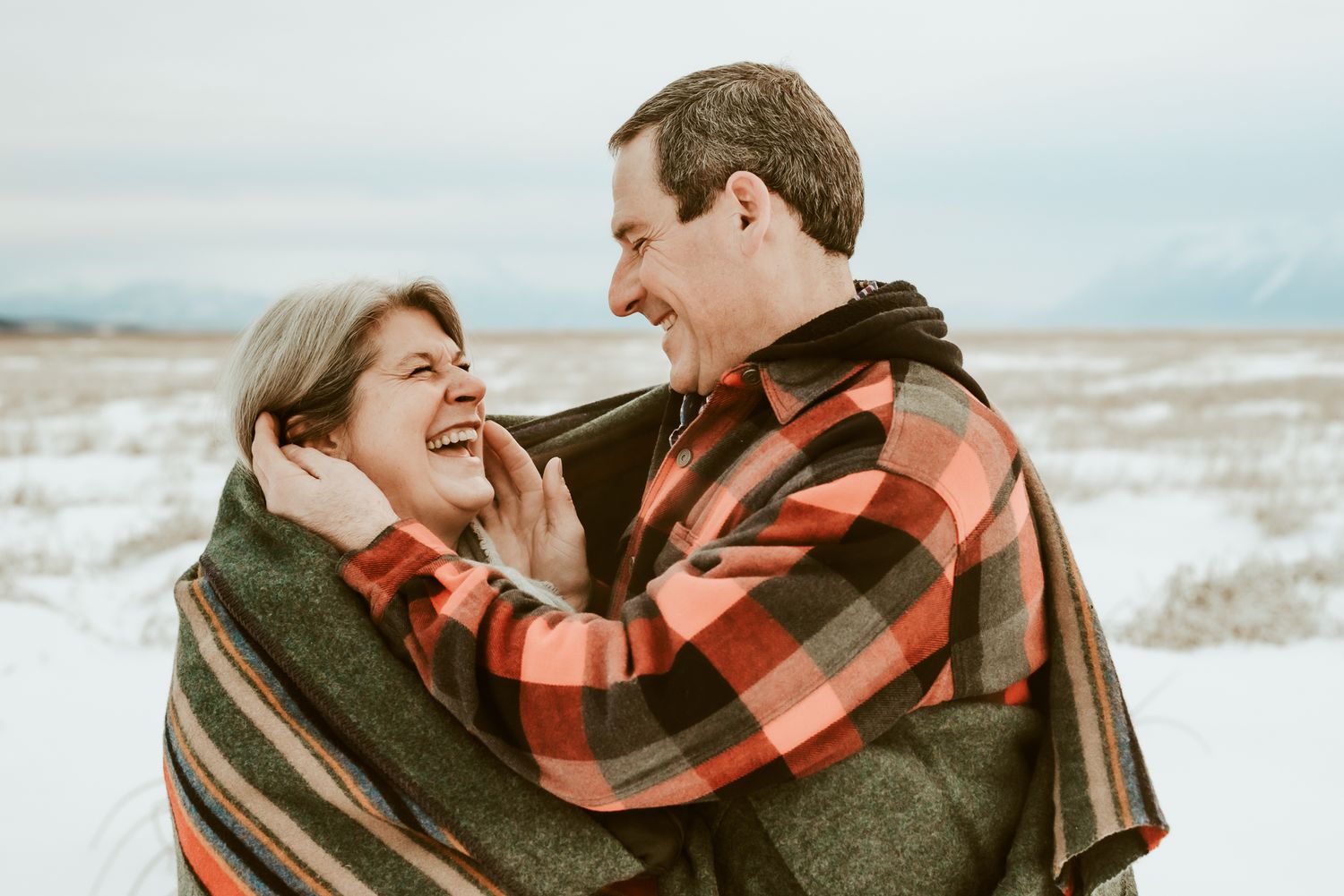 Couple wrapped in a red plaid scarf enjoying a tender moment against a snowy mountain backdrop.