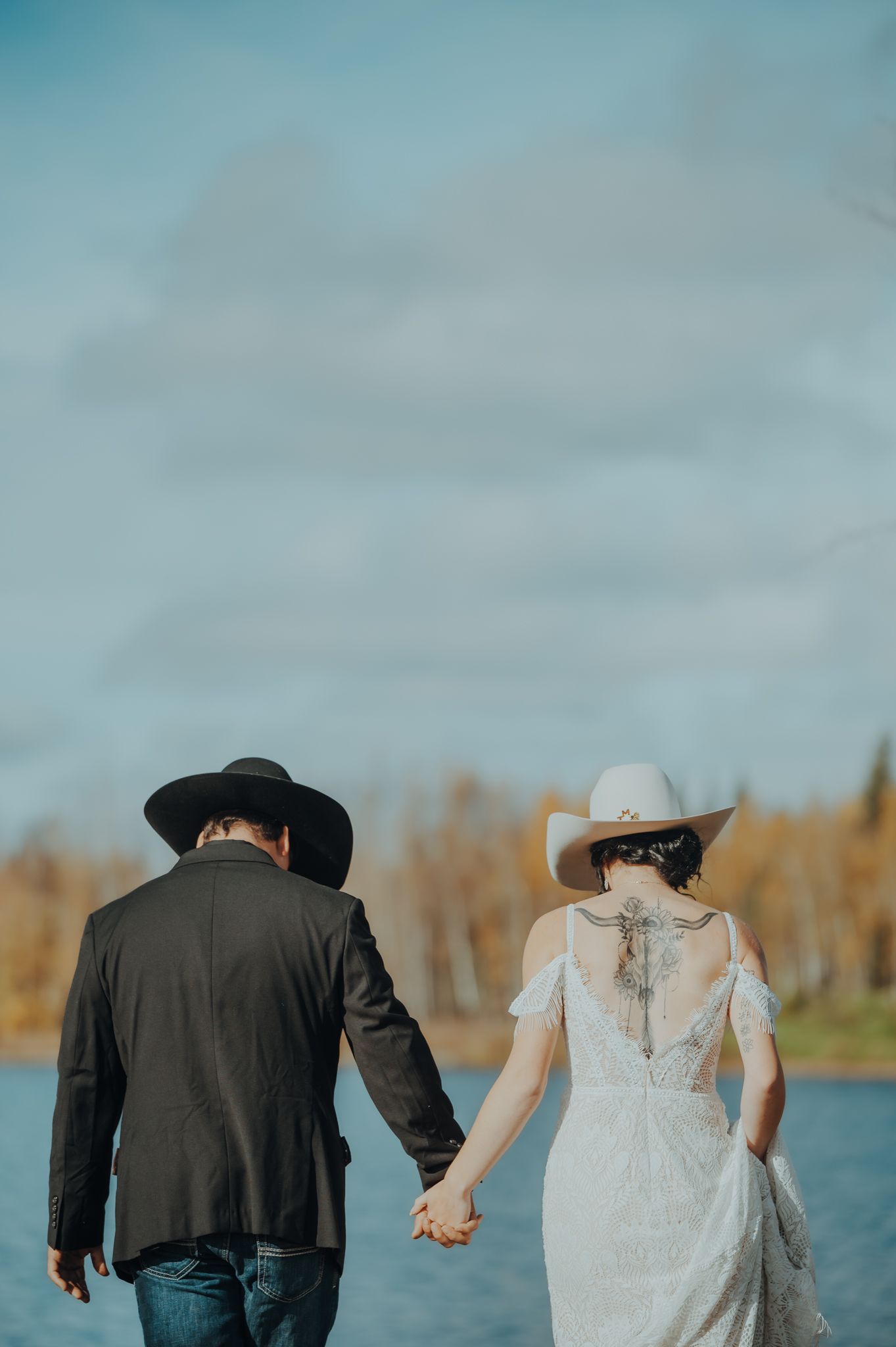 Couple in western attire walking hand in hand near a lake.