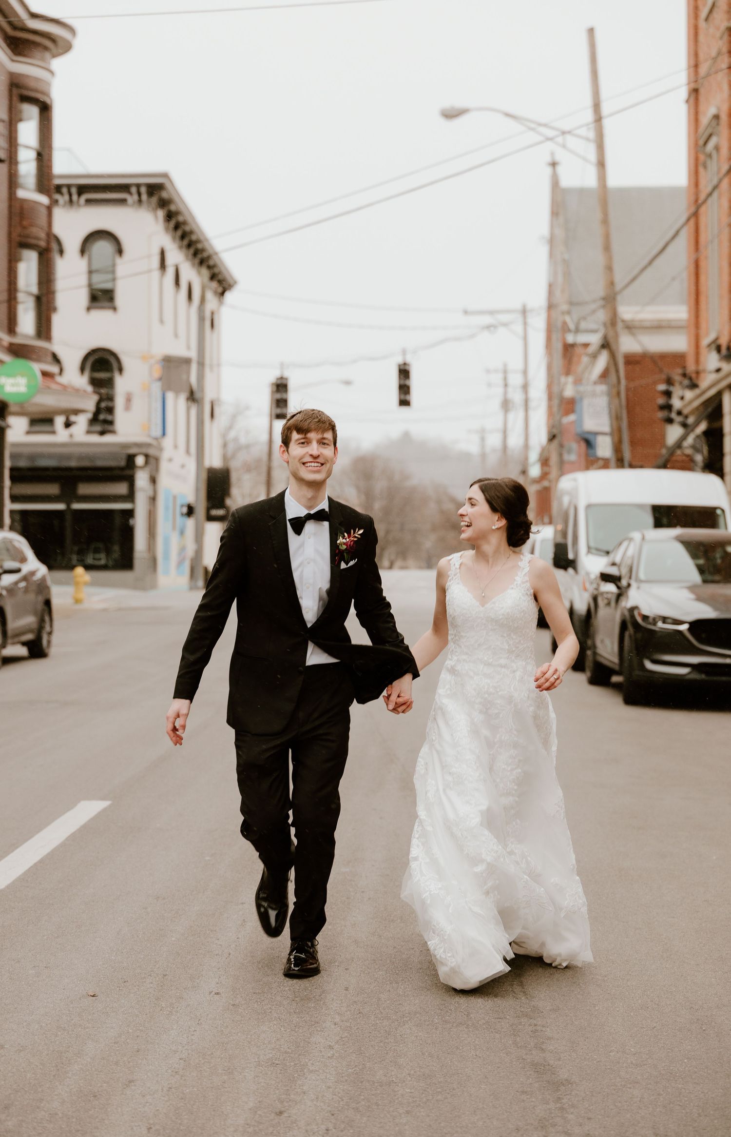 A newlywed couple runs joyfully down a city street, the bride in white and groom in black formal attire.