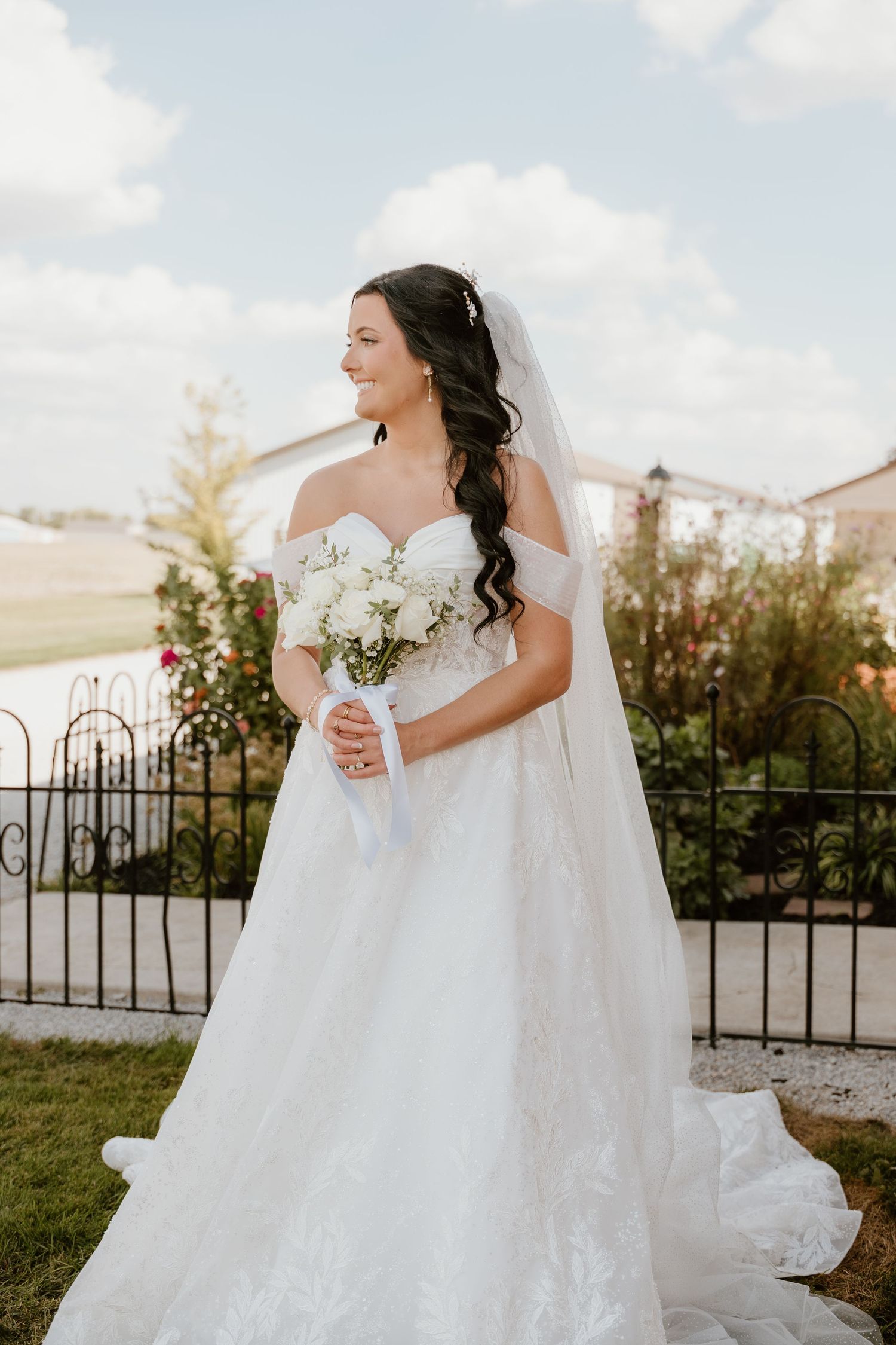 A bride in a flowing white lace wedding gown holds a bouquet while posing outdoors against an iron gate.