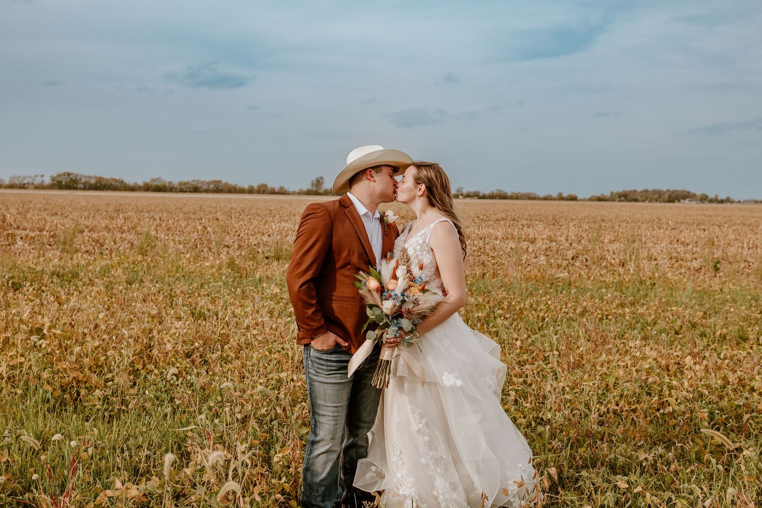 A bride and groom share a kiss in a golden field, she holds a wildflower bouquet, he wears a brown suit and white hat.