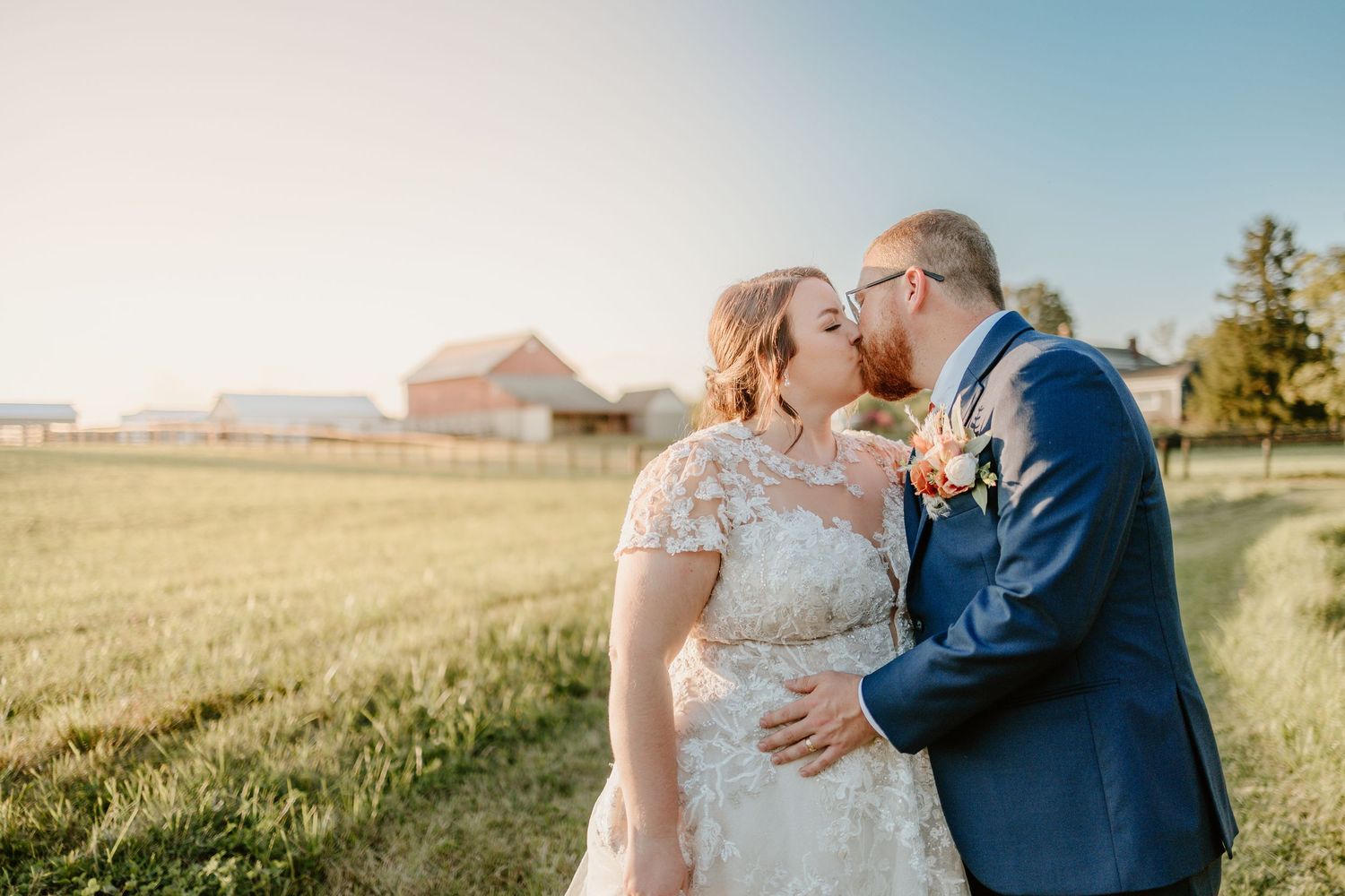 Bride and groom share a romantic kiss at golden hour in a scenic farm setting, with a red barn visible in the background.