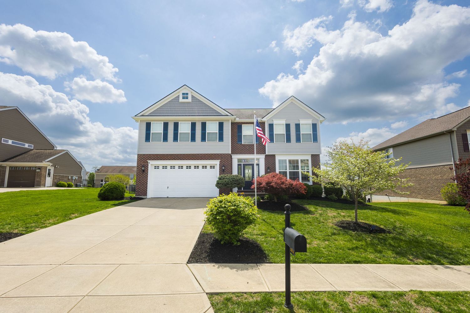 Two-story colonial home with brick accents, attached garage, manicured lawn, and American flag on a sunny day.