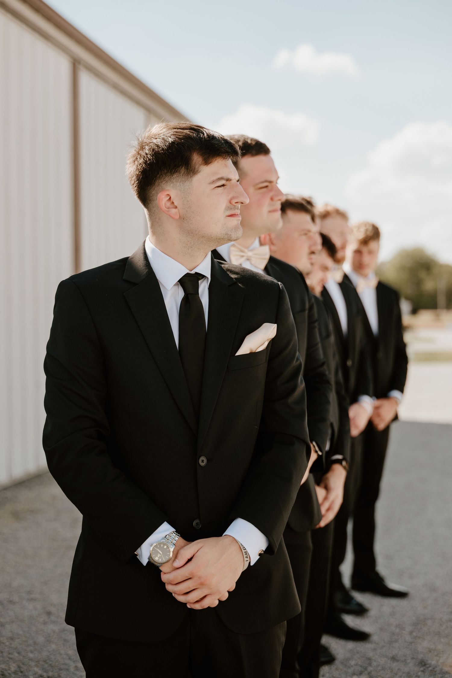 Groomsmen in black suits and ties stand in a line outside on a sunny day.