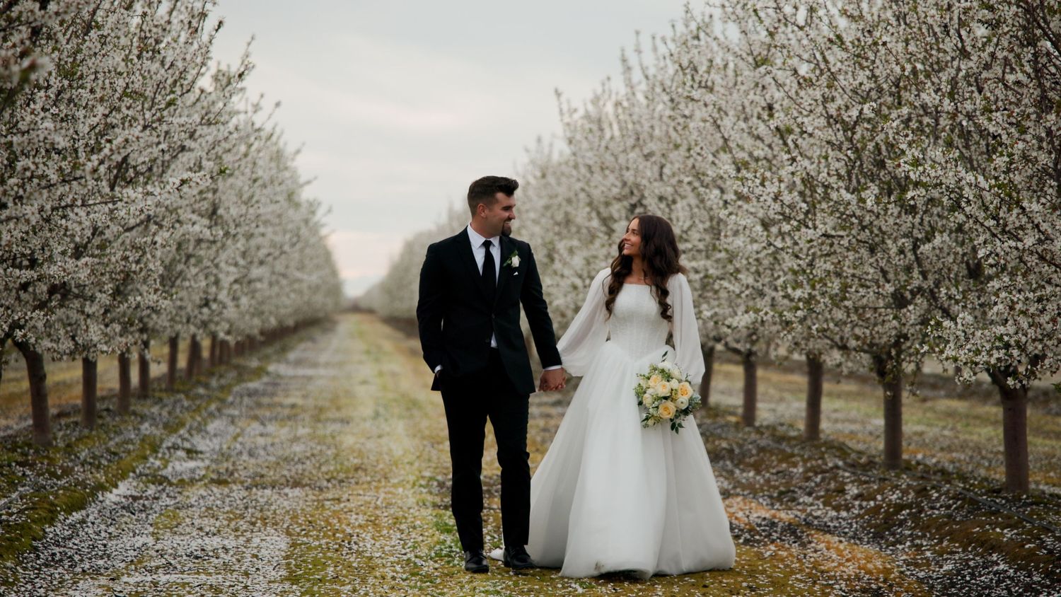 Bride and groom walk hand-in-hand through a blooming white orchard on their wedding day.