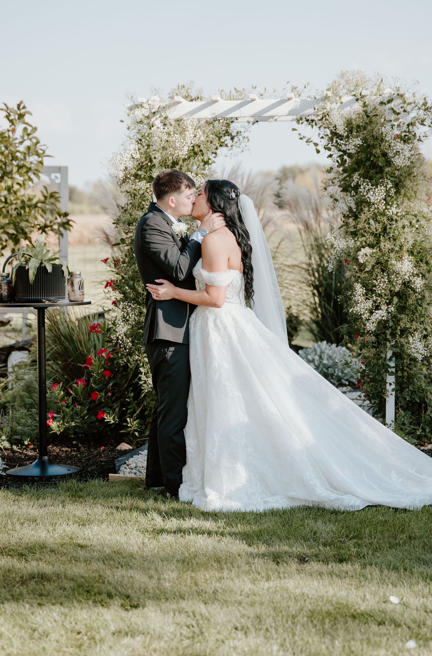 A romantic wedding couple shares their first kiss in a garden setting with lush greenery and red flowers.