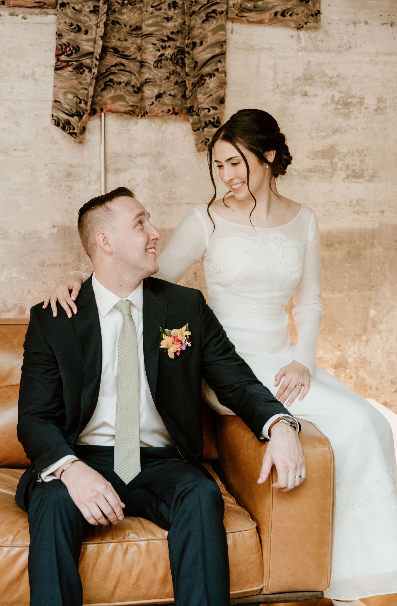 Bride in white gown sits beside groom in dark suit with floral boutonniere on leather sofa, sharing a tender moment.