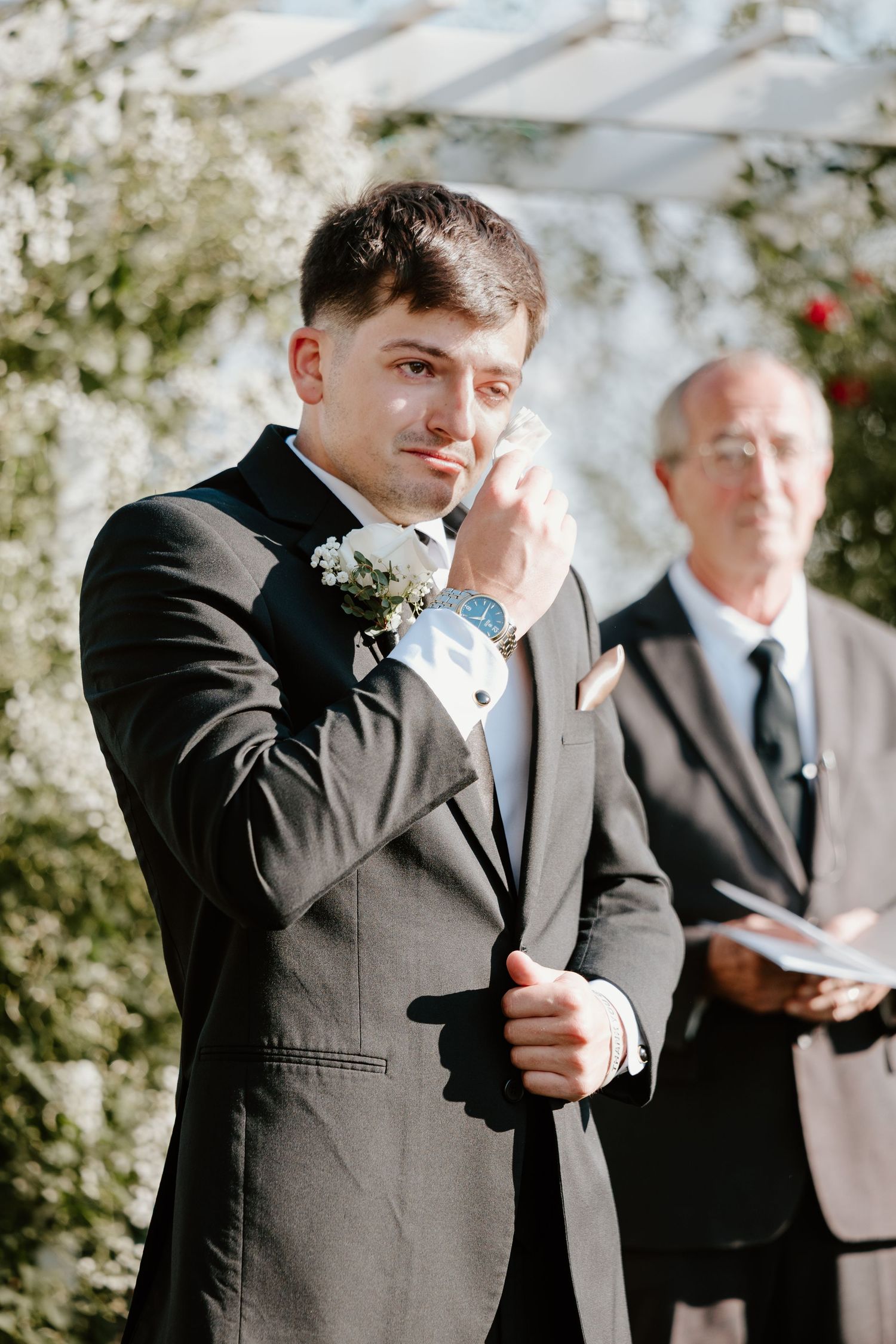 An emotional groom in a black tuxedo wipes away tears during a wedding ceremony.