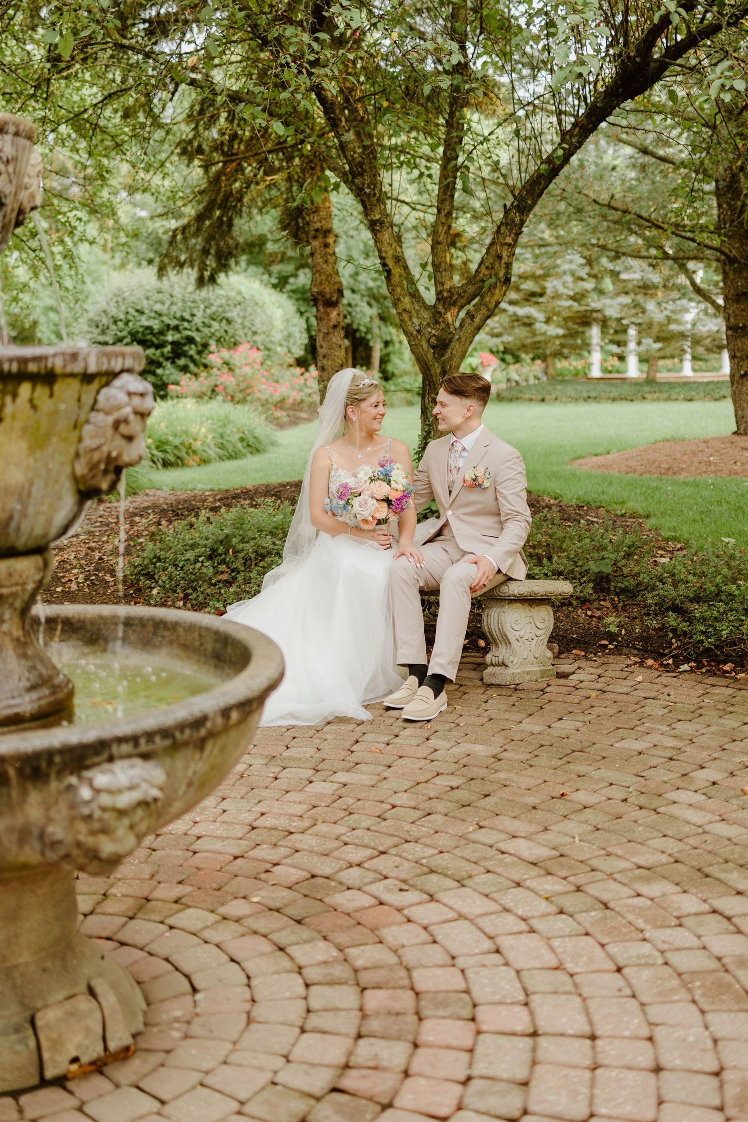 A couple sits together on a curved brick bench in a lush garden setting during a wedding photo session.