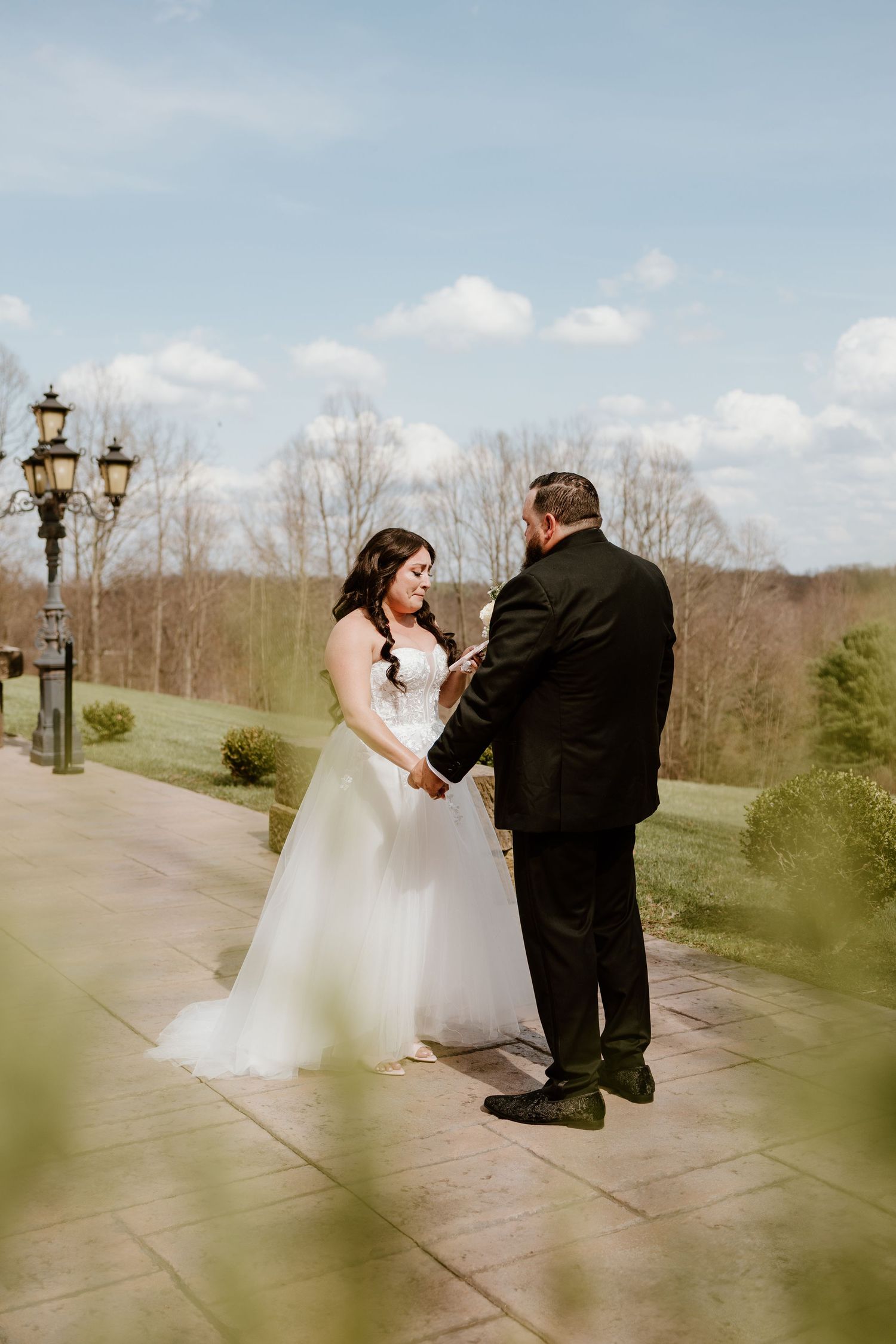 Bride and groom exchanging vows outdoors on a stone patio surrounded by lush greenery under a blue sky.