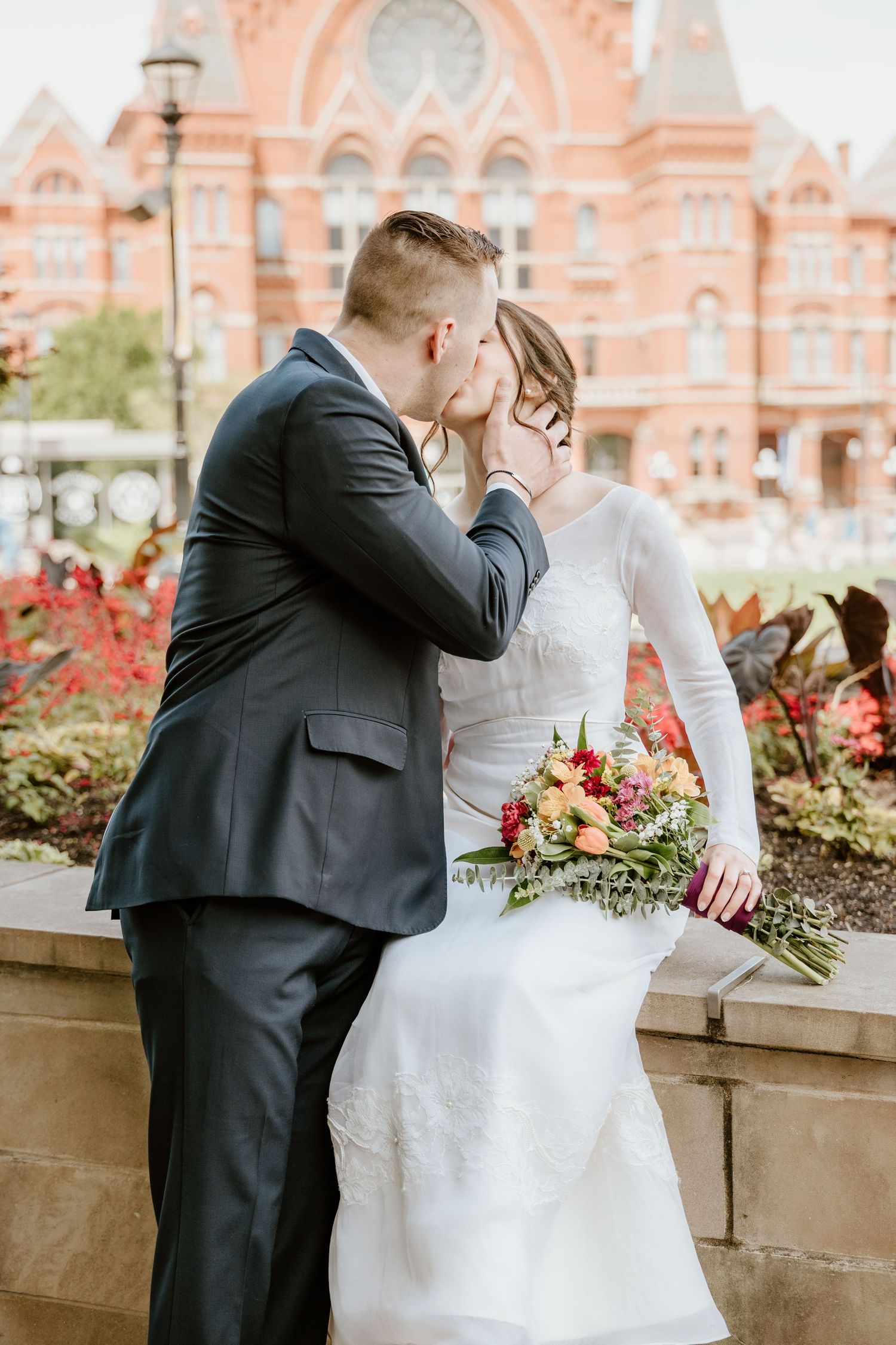 Bride and groom share a romantic kiss outdoors, with a red brick building and colorful fall flowers in the background.