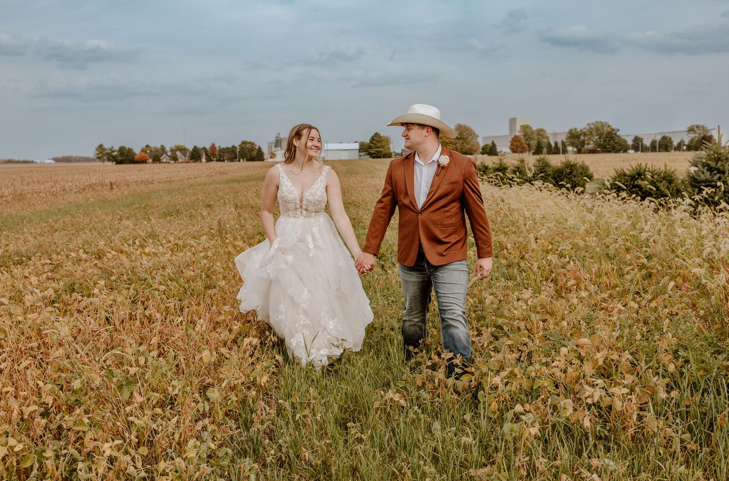 A couple walks hand in hand through a golden wheat field on a sunny day, wearing wedding attire and a cowboy hat.