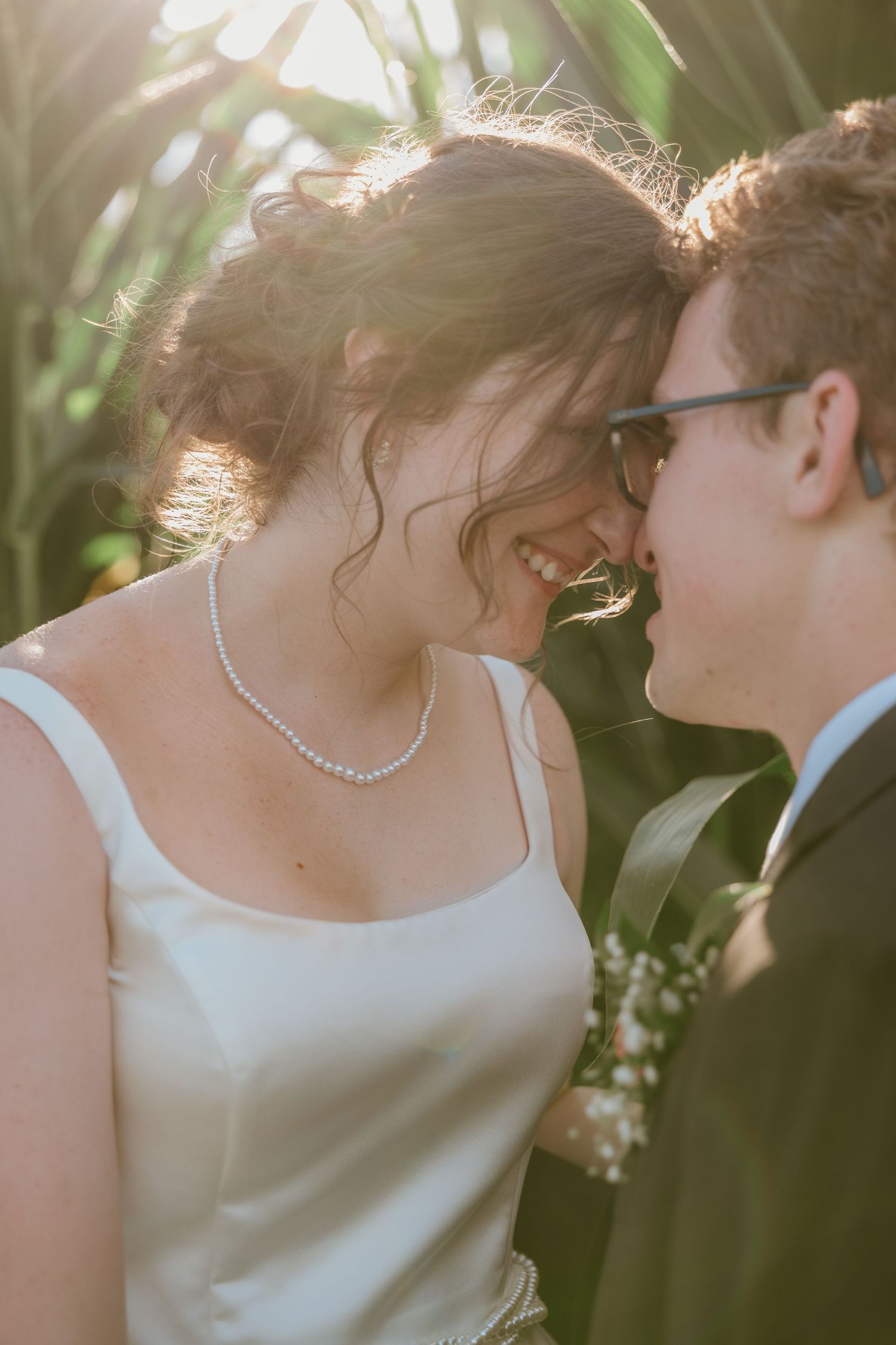 A smiling bride in a white dress and pearl necklace shares a romantic moment with a groom in glasses outdoors.