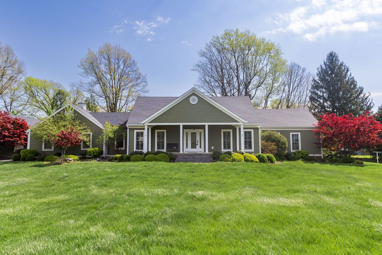 A charming green ranch-style home with white trim, lush lawn, and colorful flowering trees on a sunny spring day.