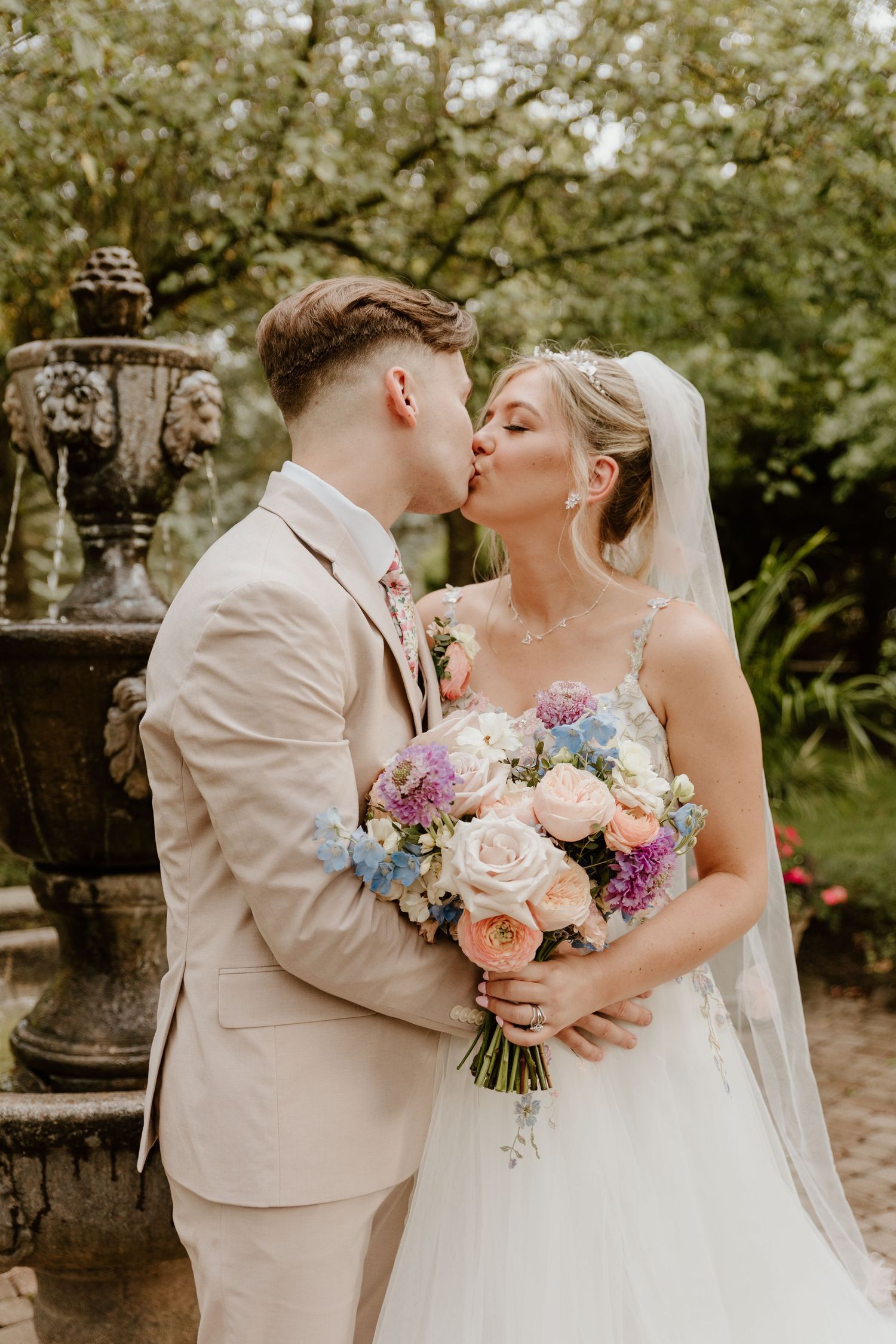 A romantic wedding couple shares intimate moments in a garden setting with pastel floral bouquets and light-colored attire.